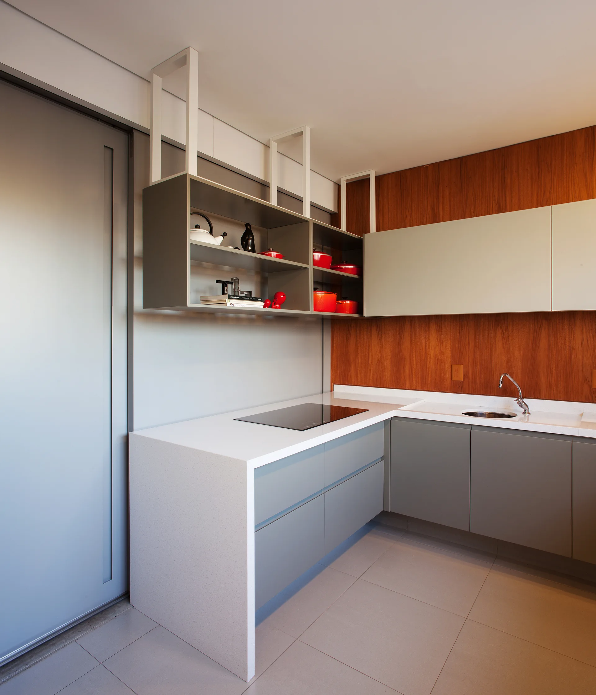 A modern kitchen featuring grey flat-panel cabinets, a white waterfall countertop, ceiling-suspended open shelving, and a wood-paneled accent wall.