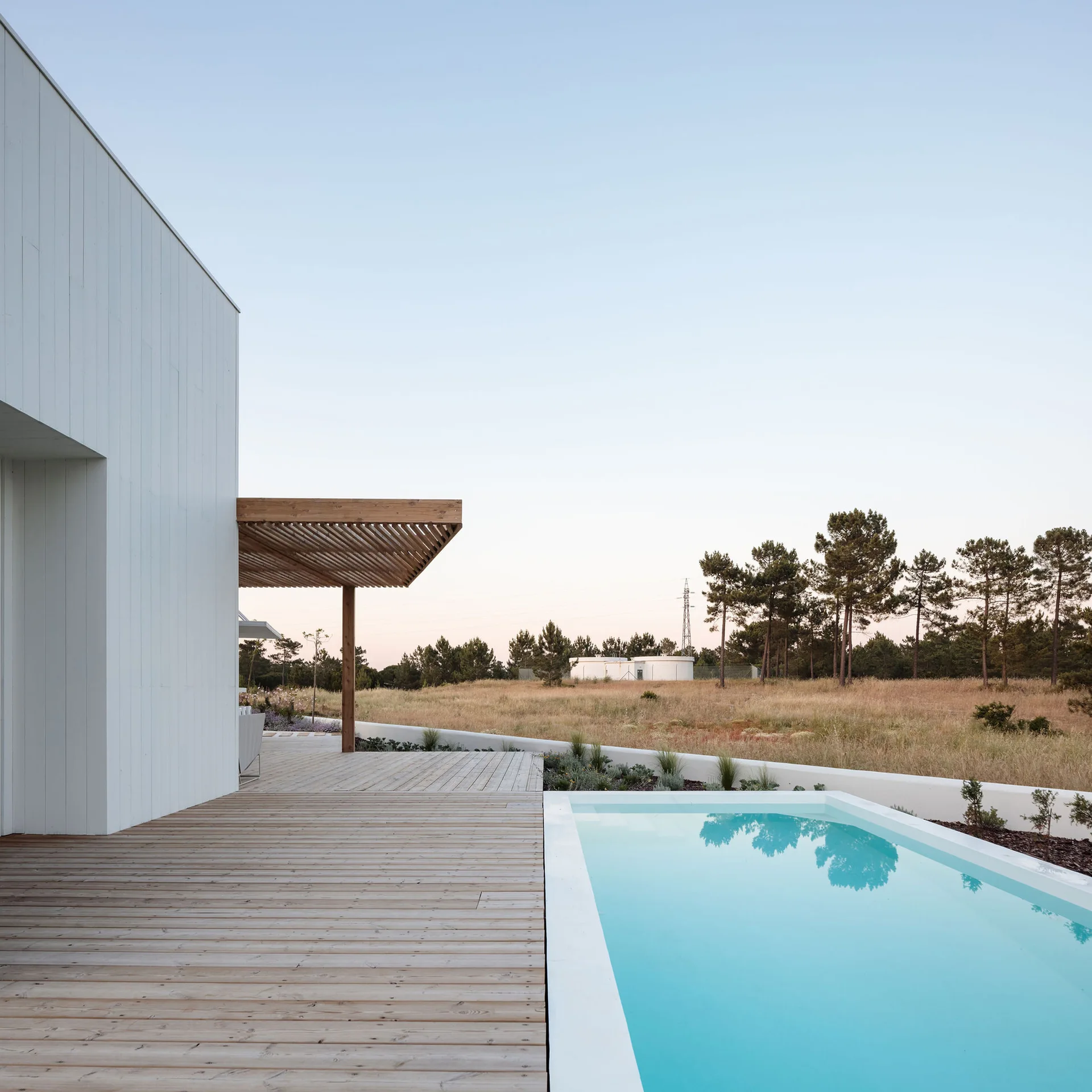 A rectangular swimming pool sits next to a light wood deck and a modern white-clad house with a slatted wooden pergola.