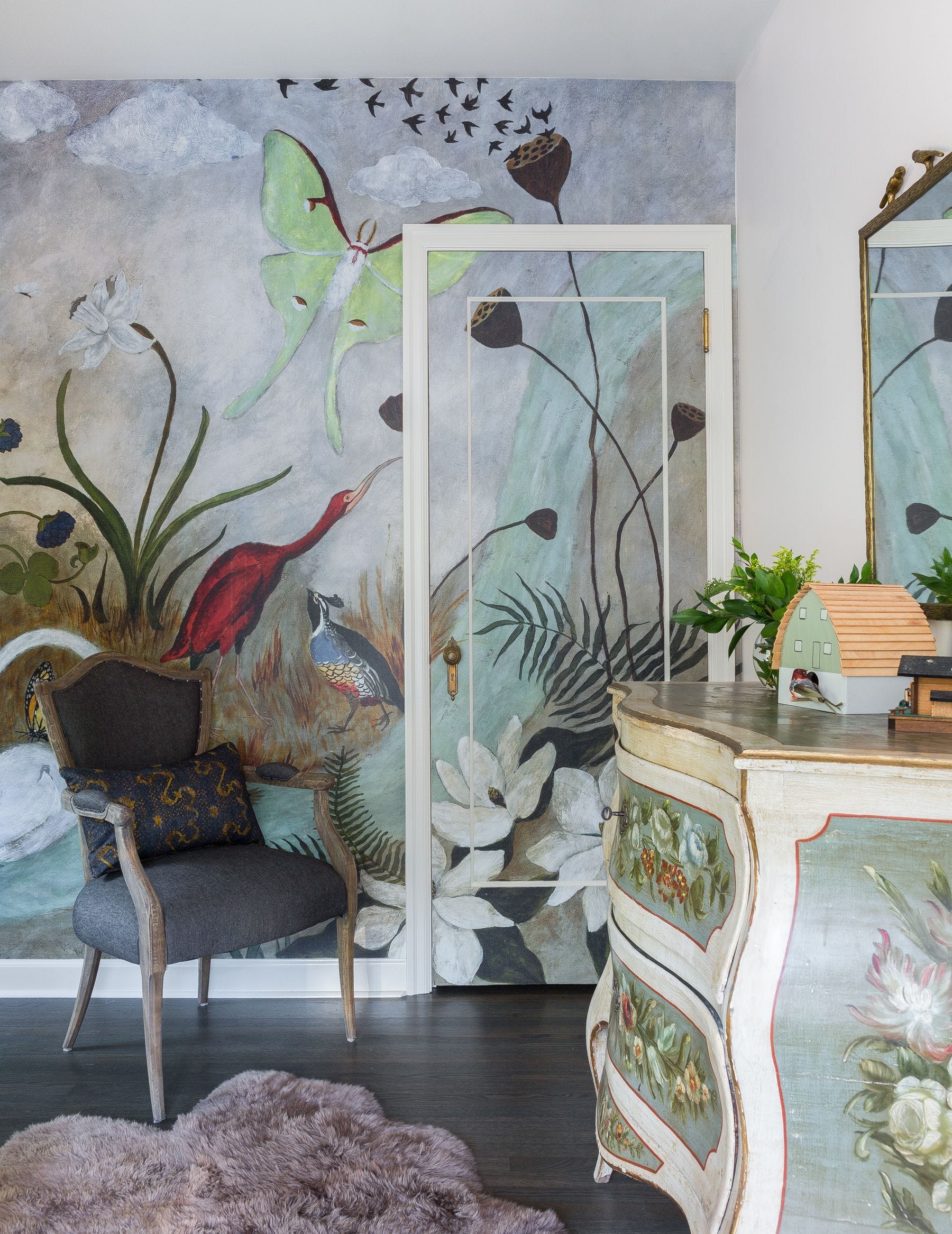 A nature-themed mural featuring birds and flowers extends across a wall and integrated door next to a dark grey armchair and painted dresser.