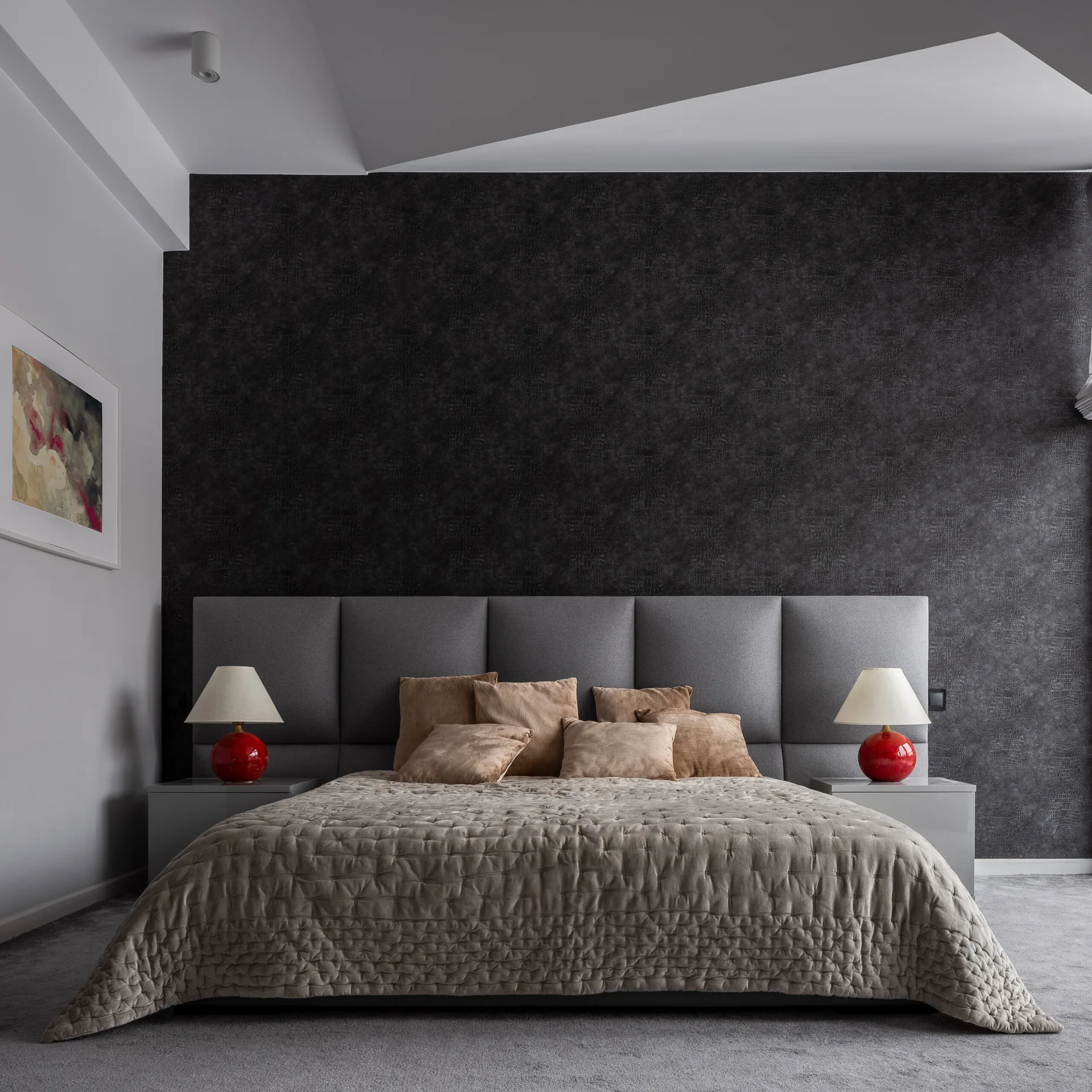 A modern bedroom with a wide gray padded headboard against a dark textured accent wall, featuring neutral bedding and two glossy red bedside lamps.