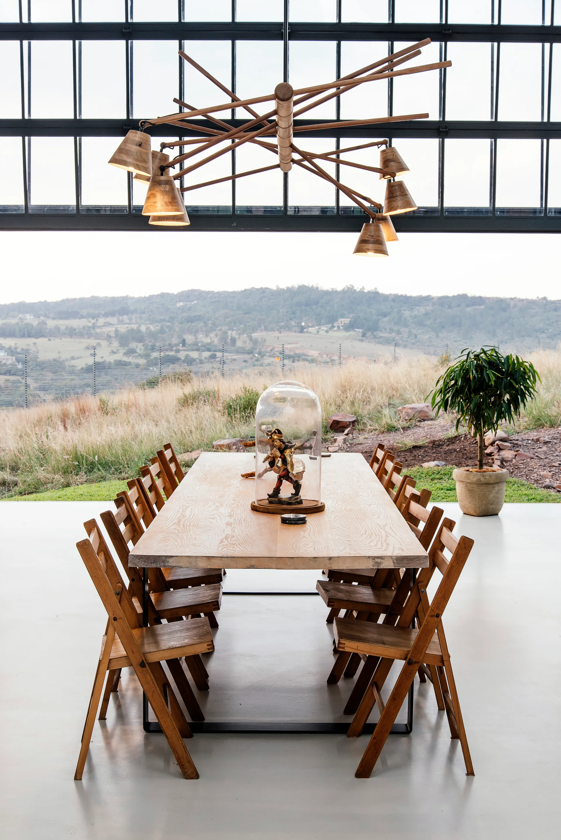 A long wooden dining table surrounded by matching chairs is set before a large gridded window overlooking a grassy hill.