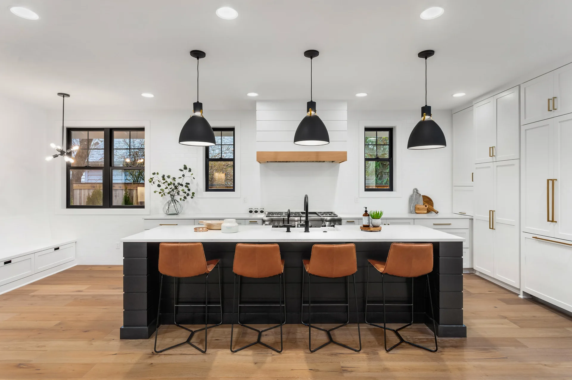 A bright kitchen featuring a black shiplap island with four tan leather barstools, black pendant lights, and white perimeter cabinetry with brass hardware.
