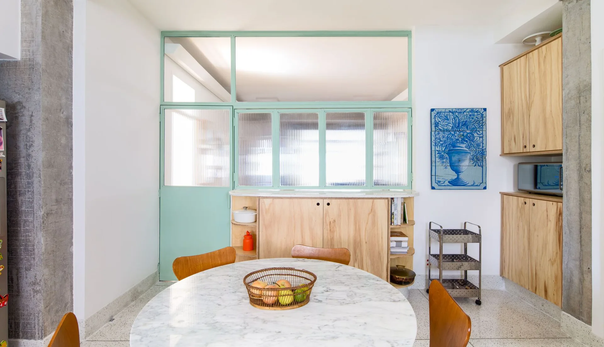 A dining space features a round marble table, light wood cabinetry, a raw concrete column, and a mint green glass partition wall.