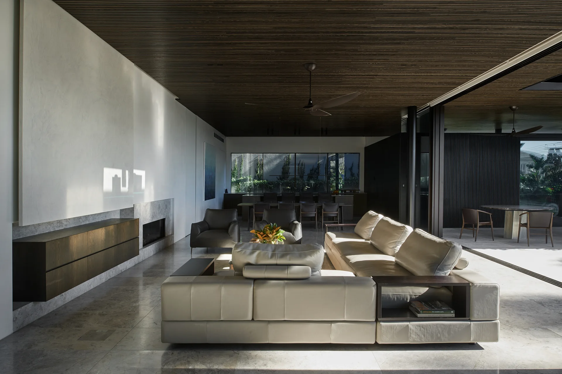 A modern open-plan living room features a cream leather sectional sofa, a slatted dark wood ceiling, and sliding glass doors leading to an outdoor patio.