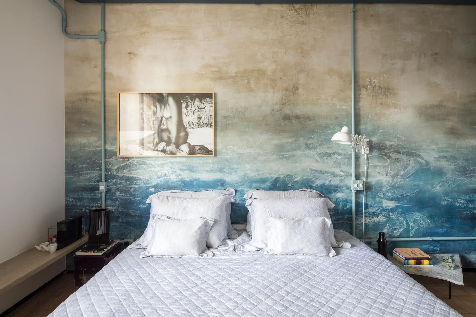 A bedroom featuring a double bed with quilted bedding set against a blue and beige ombre wall mural with exposed painted conduit.