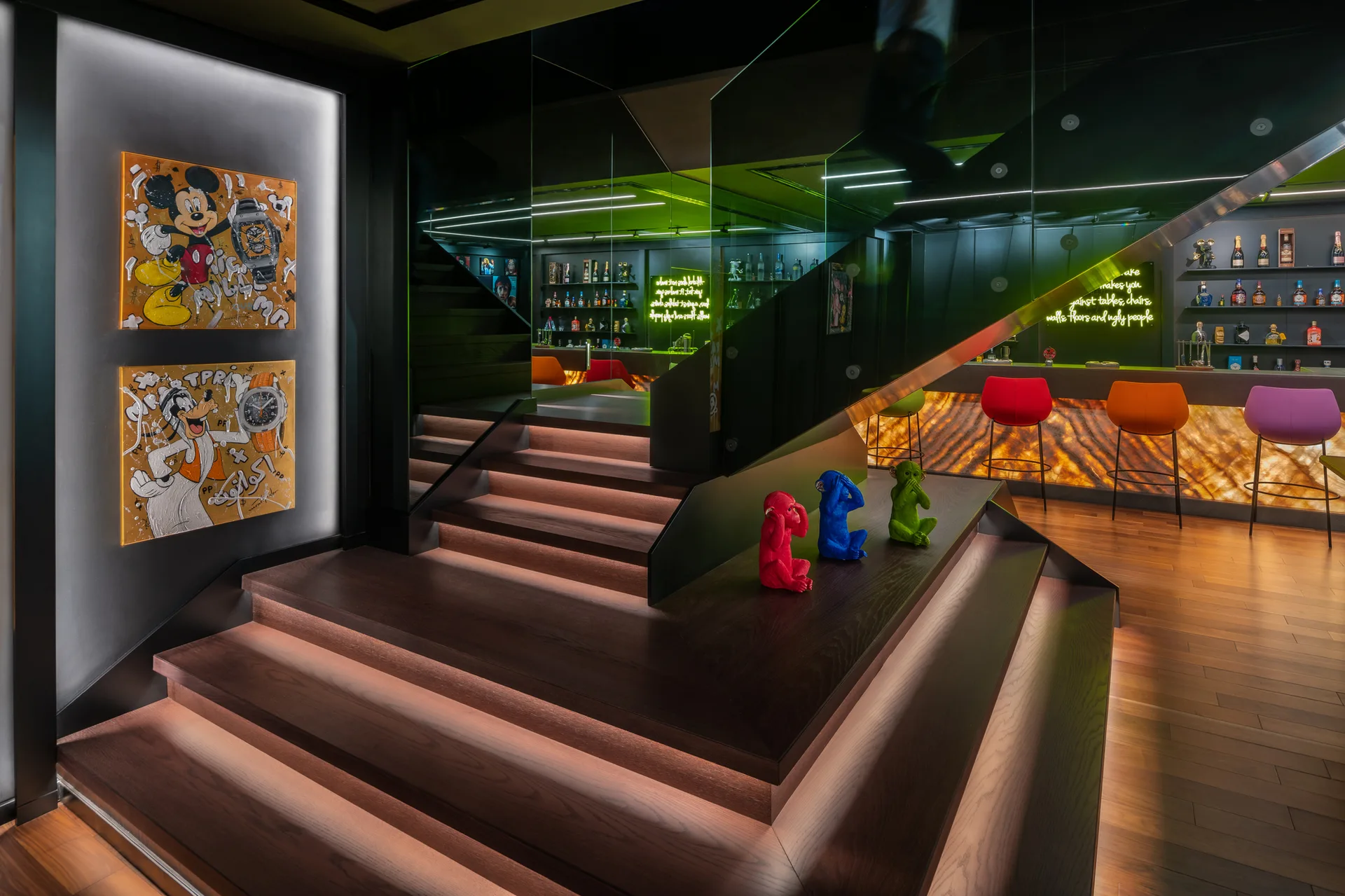 A dark wood staircase with glowing step treads stands next to a backlit lounge bar featuring colorful stools and neon wall signs.
