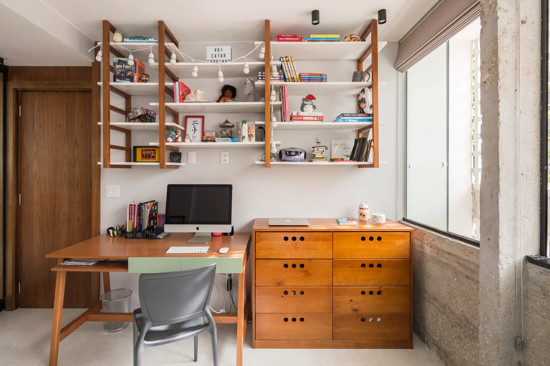 A wooden desk and matching dresser sit below open shelves in a home office next to a concrete pillar.