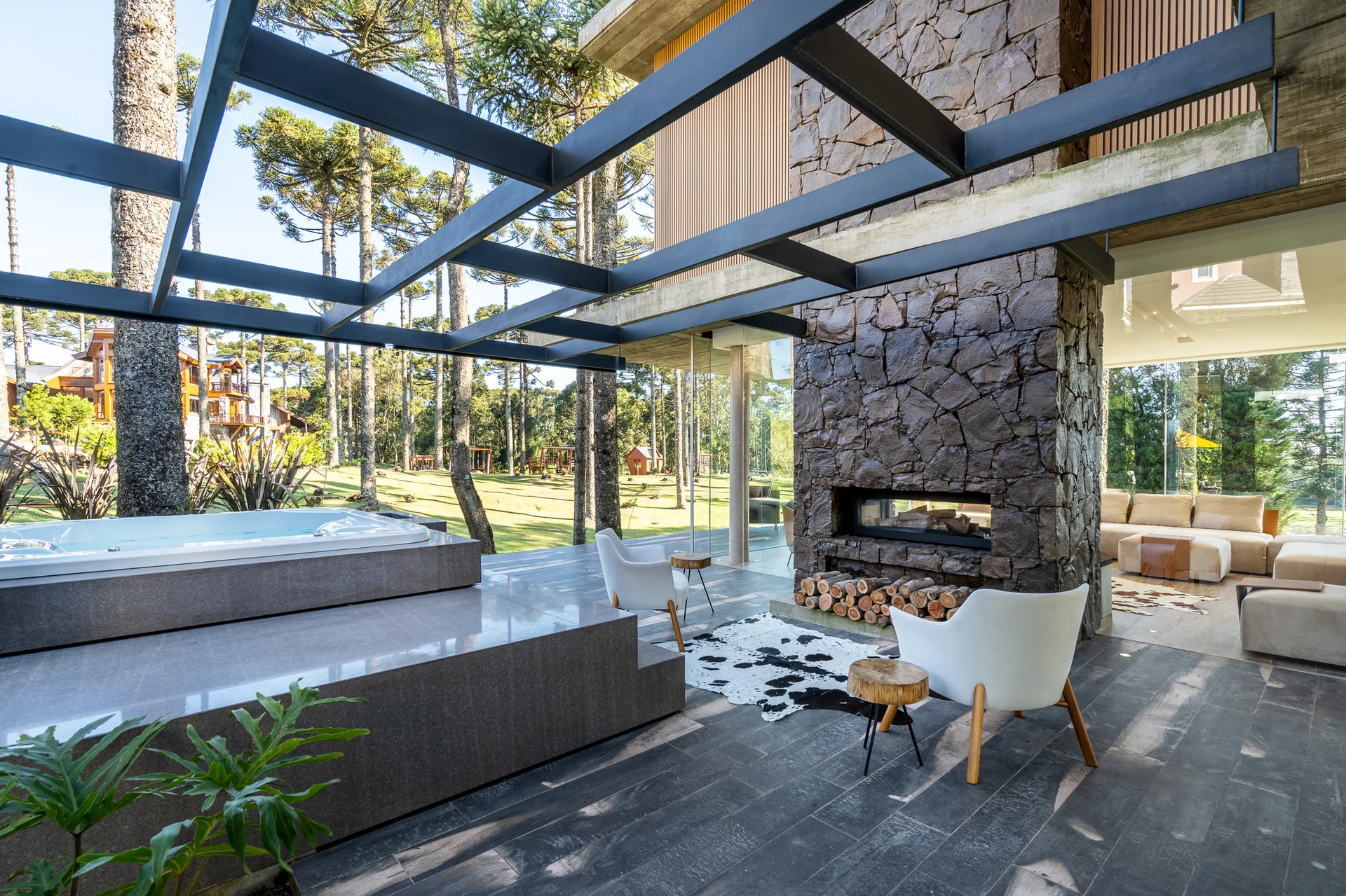 A modern outdoor patio features a stone-clad fireplace, two white armchairs on a cowhide rug, a built-in hot tub, and a black metal pergola.