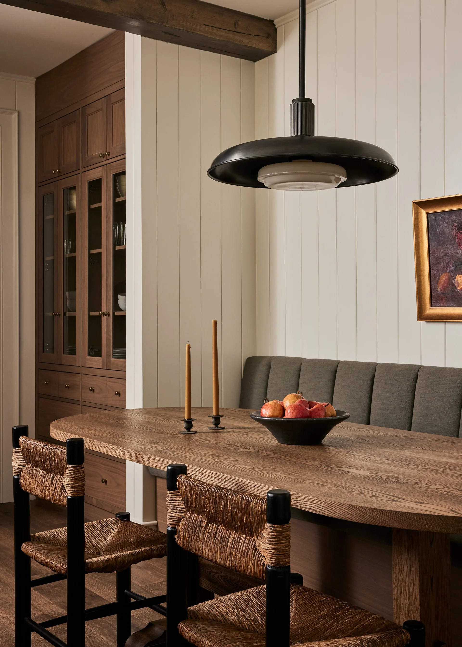 A dining area features a wood oval table, black chairs with woven seats, a channel-tufted banquette, and a large black pendant light.