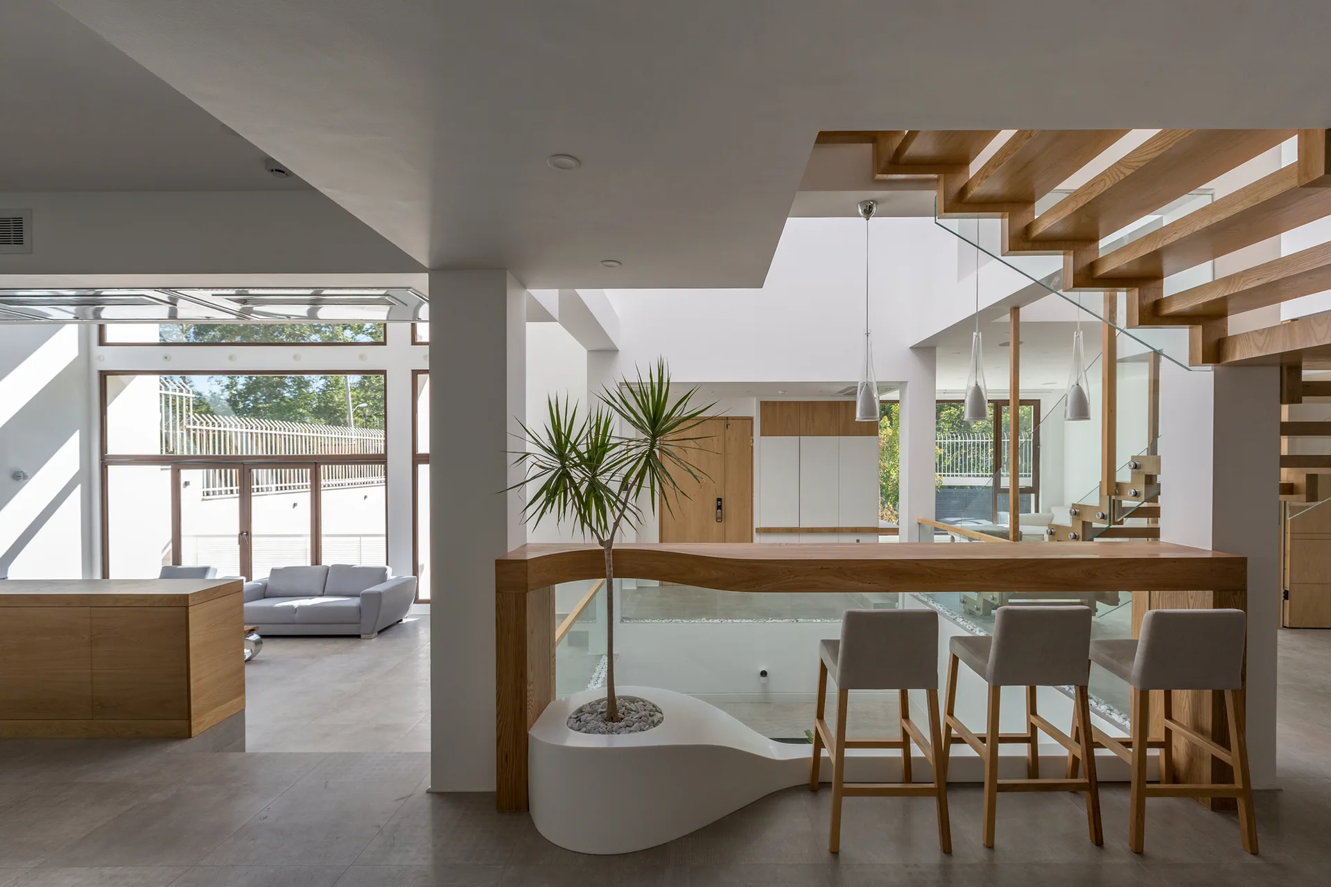 A modern open-concept home interior featuring a light wood bar counter with gray bar stools, a white sculptural planter, and a floating wooden staircase.