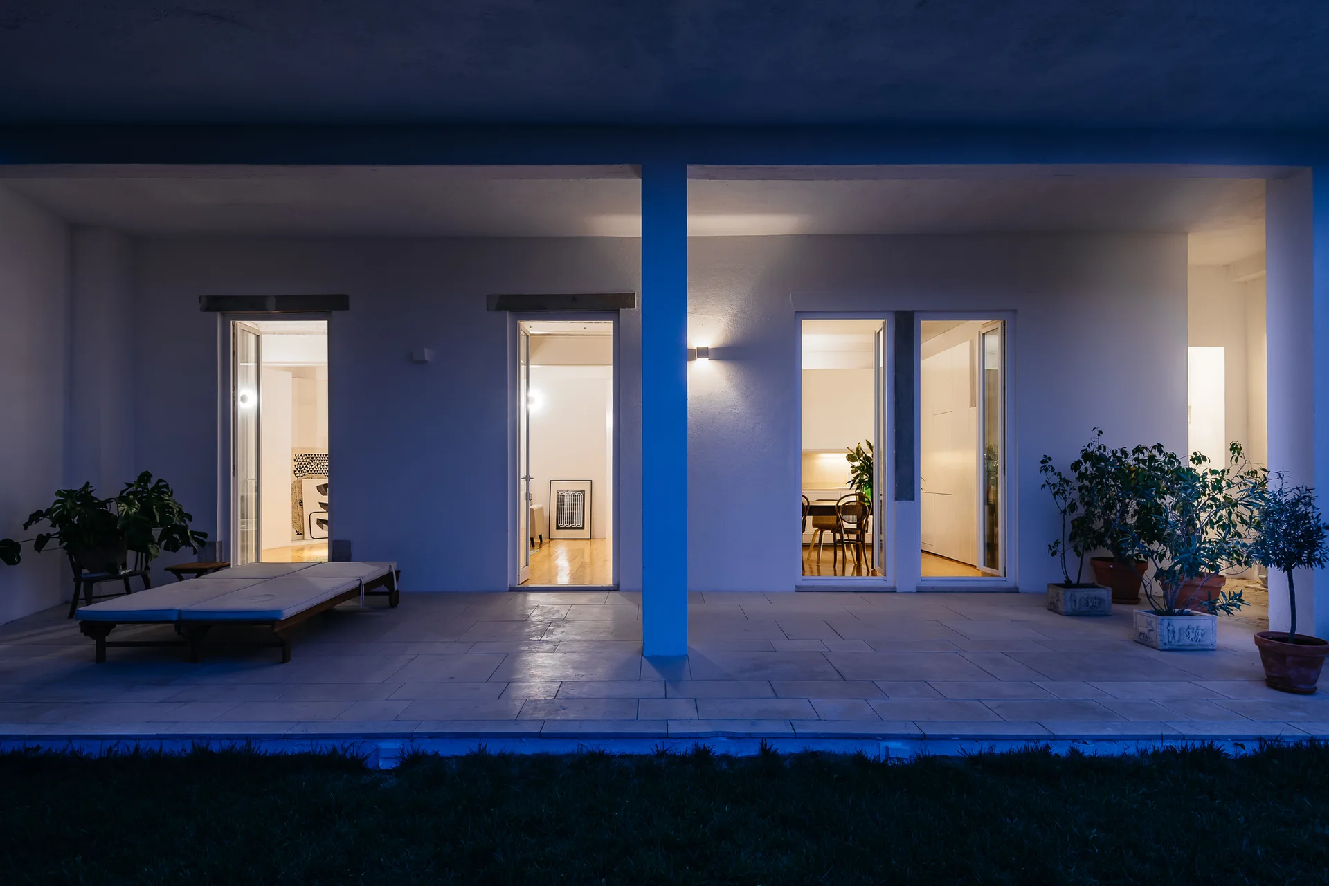 A covered patio with stone tiles features a wooden daybed and potted plants outside glass doors glowing with warm interior light at twilight.