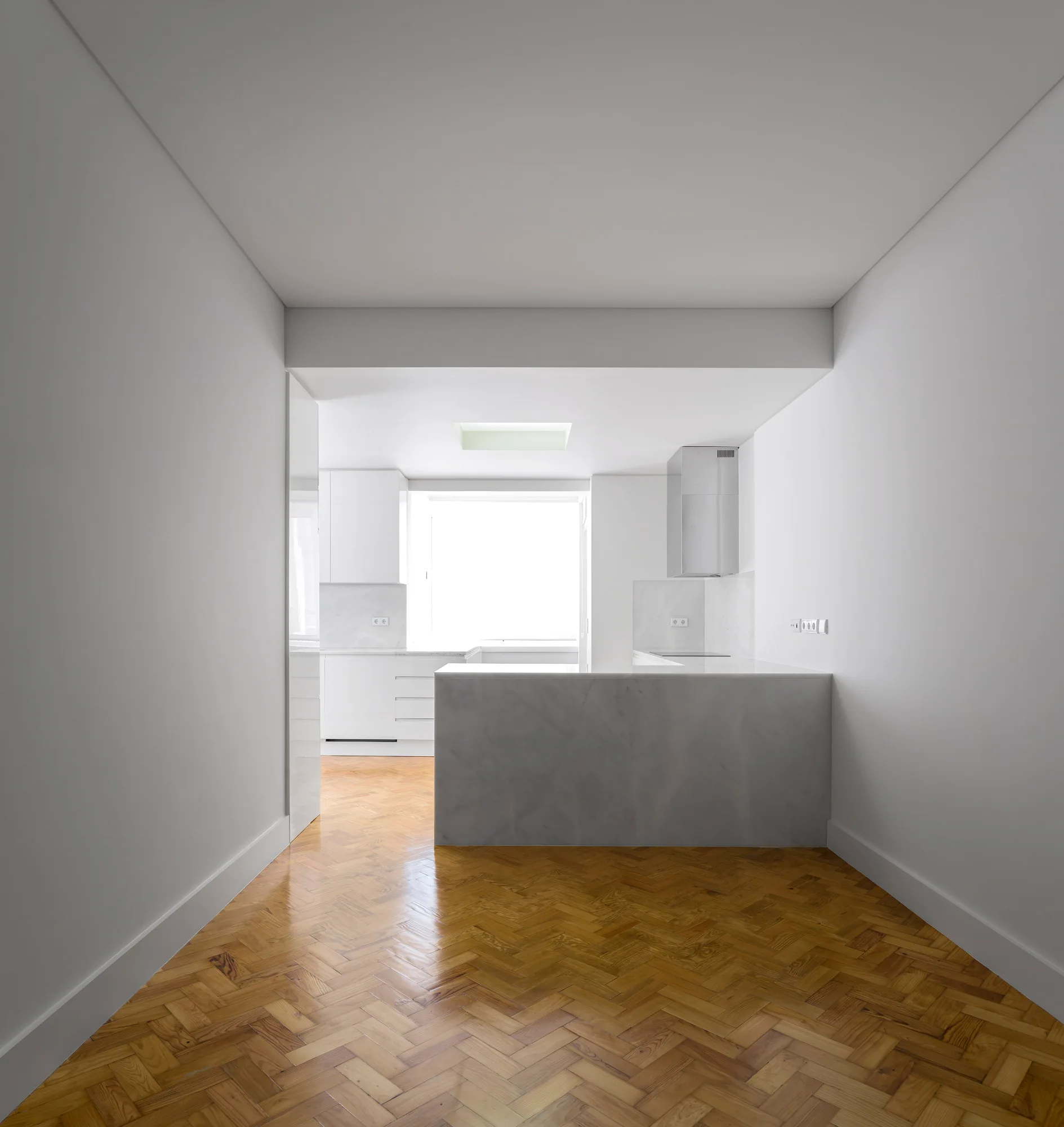 A minimalist white kitchen features a gray stone island and warm herringbone wood parquet flooring leading toward a bright window.