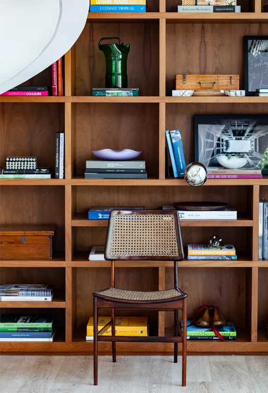 A dark wood chair with a woven cane back stands in front of a warm wood built-in shelving unit filled with books and decorative art.
