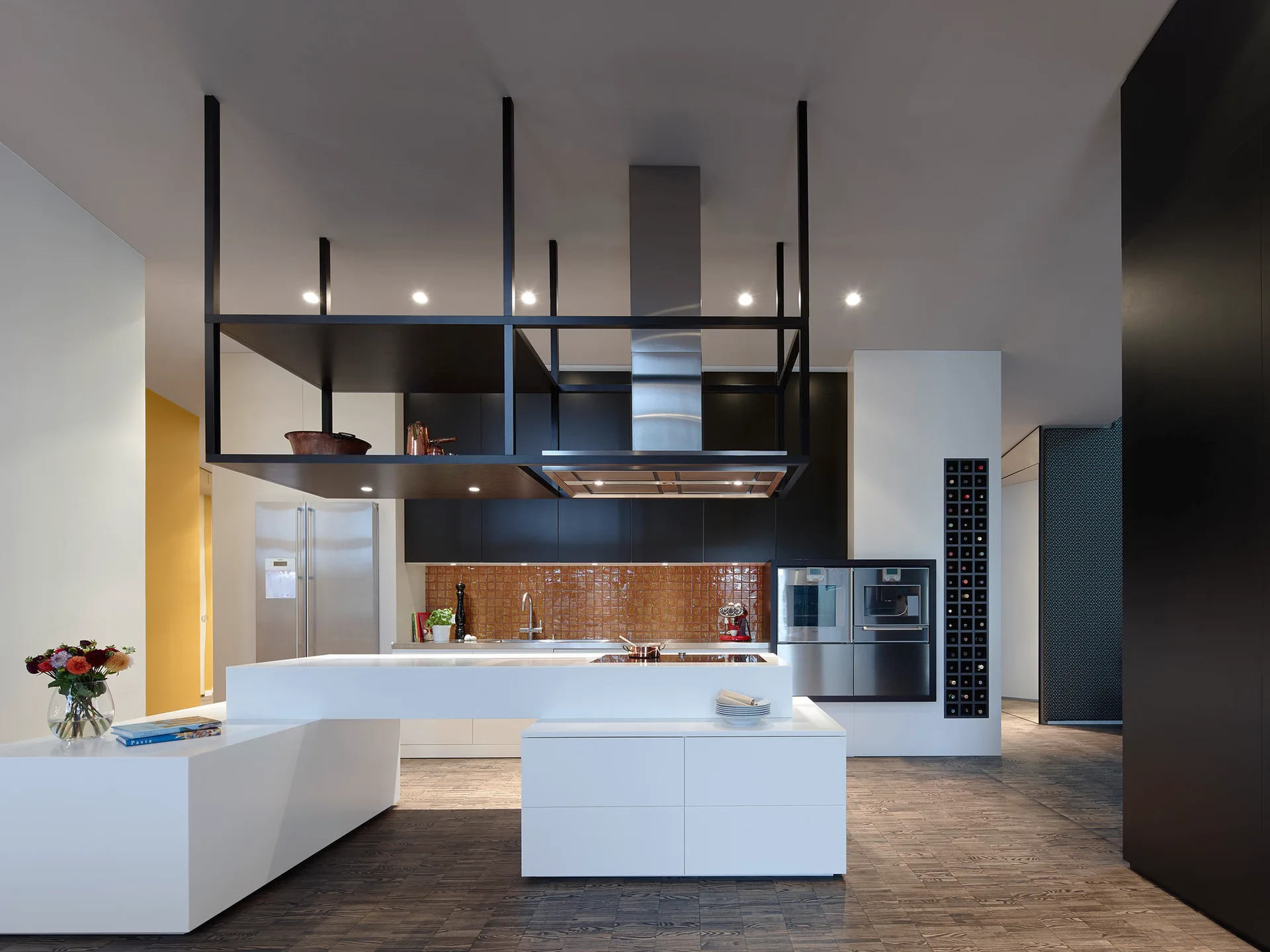 A modern kitchen featuring a multi-level white island, a black suspended ceiling shelf, and a brown square-tiled backsplash.