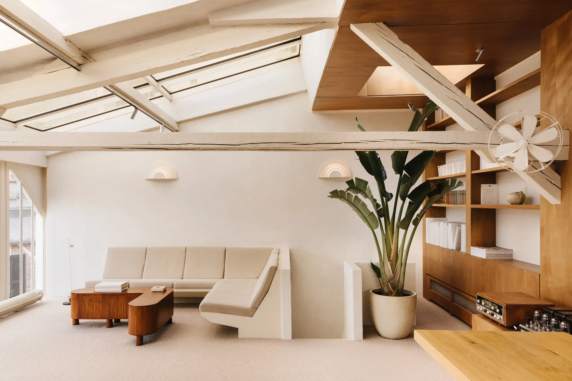 An attic living room features a cream sectional sofa, curved wooden coffee table, built-in library shelves, and exposed ceiling beams under a skylight.