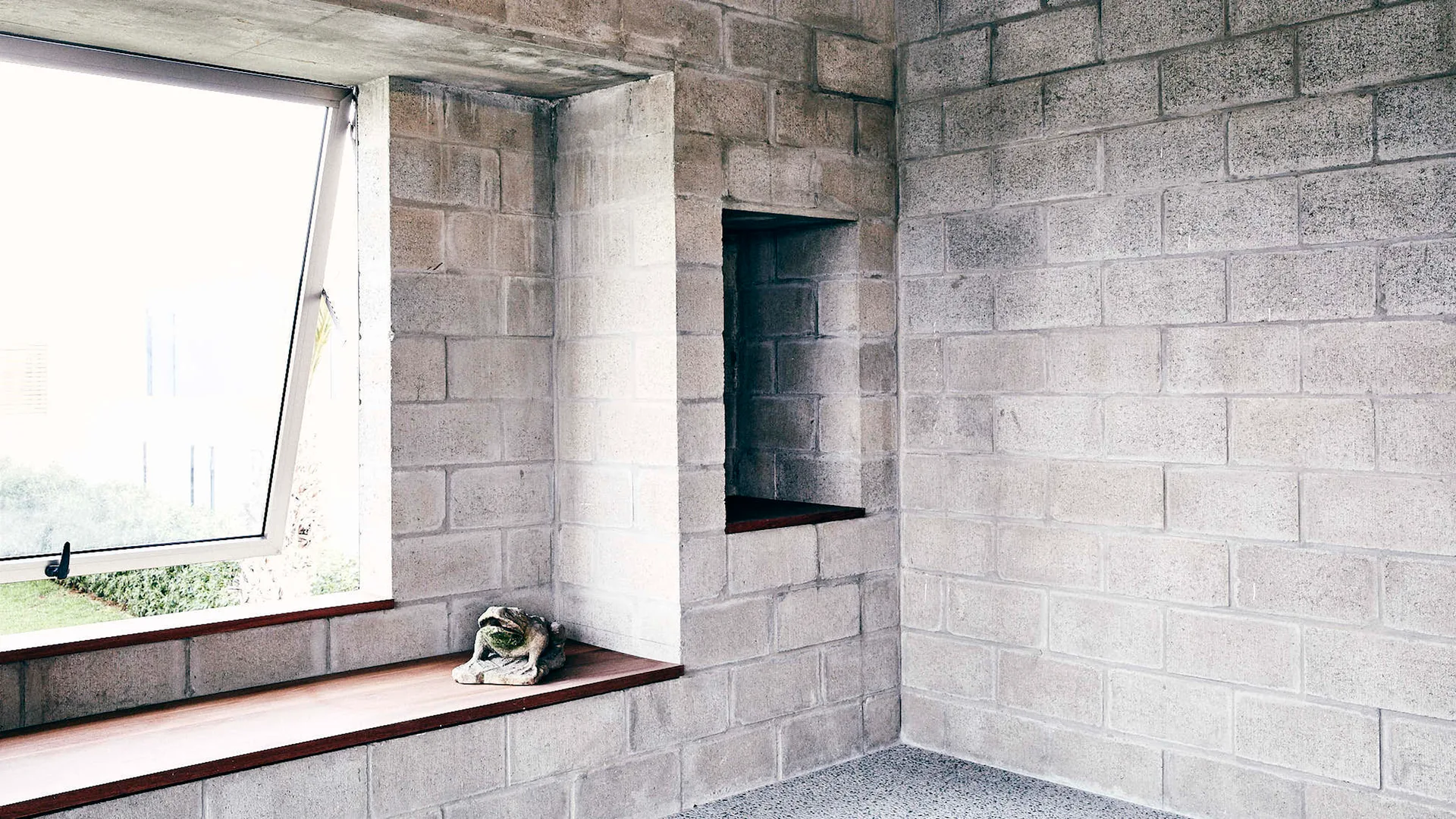 An interior with raw light grey concrete block walls, a large window with a deep wood sill, and a matching recessed wall niche.