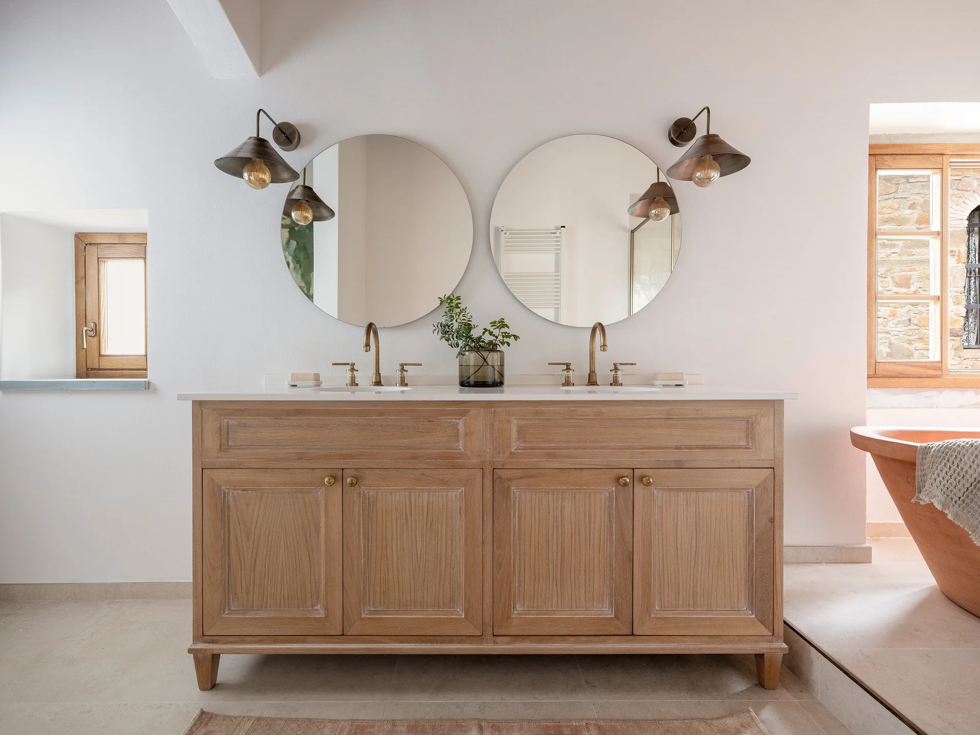 A wooden double vanity with two round mirrors, brass faucets, and dark metal wall sconces against a white plaster wall.