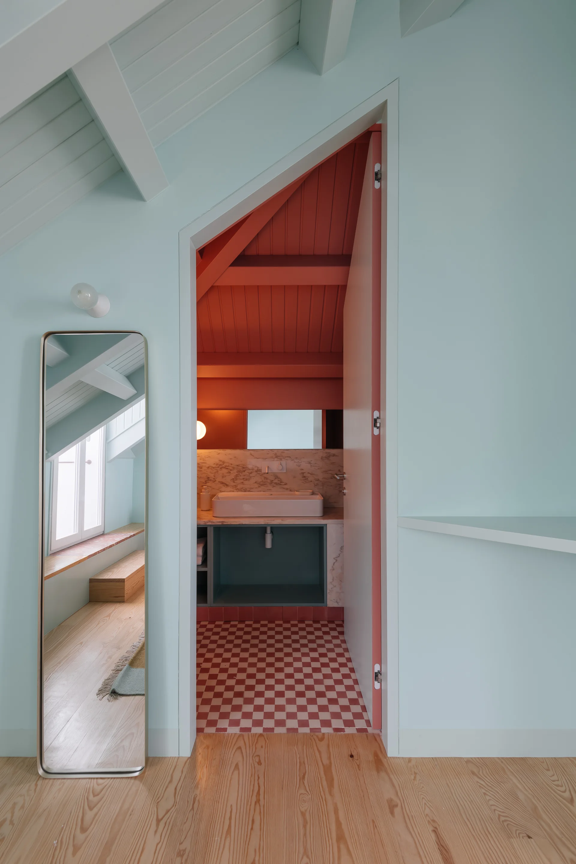 A view through a doorway into a terracotta-painted attic bathroom with checkered tile floors, contrasting with mint green walls and a floor mirror outside.
