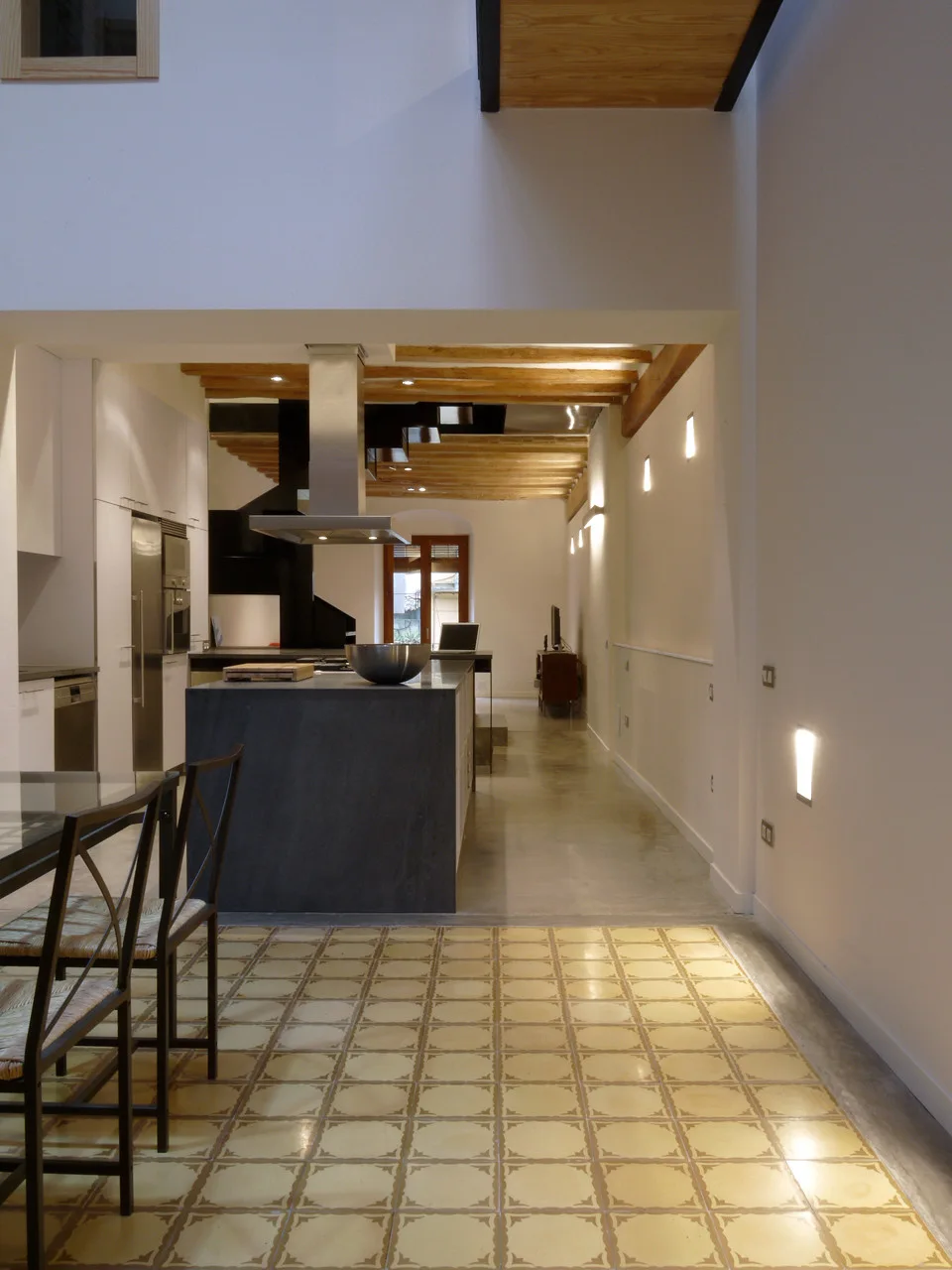 A modern open kitchen featuring a dark grey central island, white flat-front cabinets, yellow patterned floor tiles, and exposed wood ceiling beams.