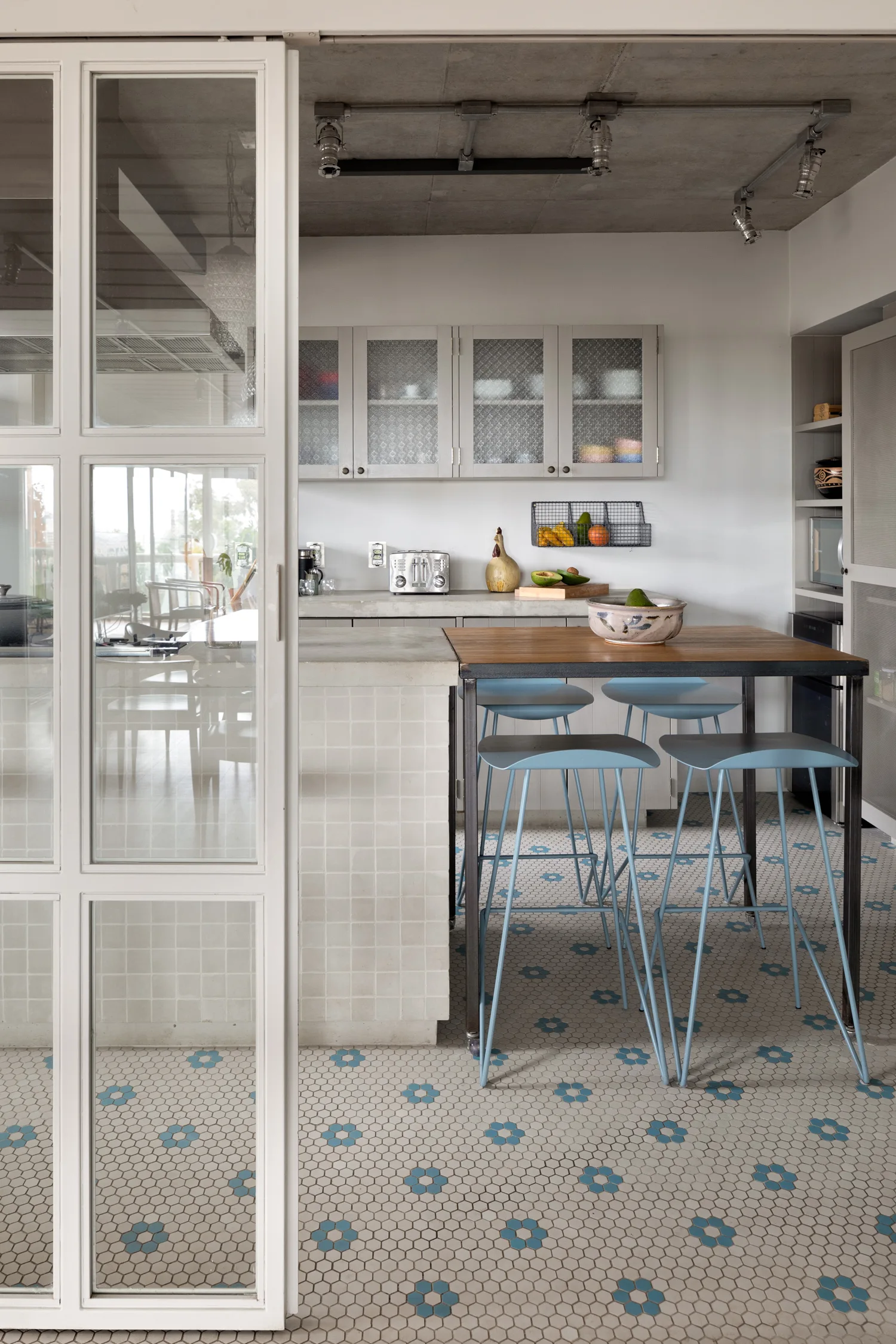 A modern kitchen features a concrete island with blue bar stools, a wooden table extension, and a flower pattern hexagonal mosaic tile floor.