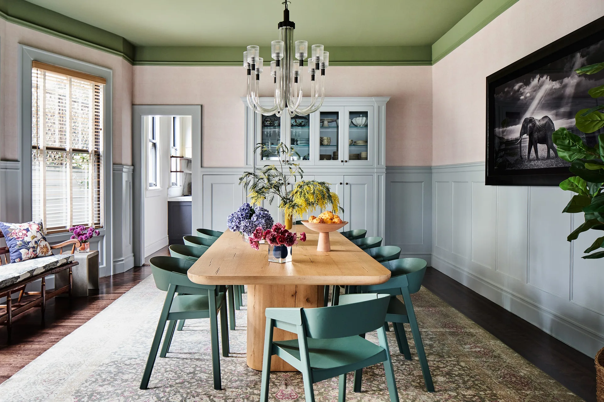 A dining room features a natural wood table surrounded by green chairs, set against gray wainscoting, blush pink walls, and a sage green ceiling.