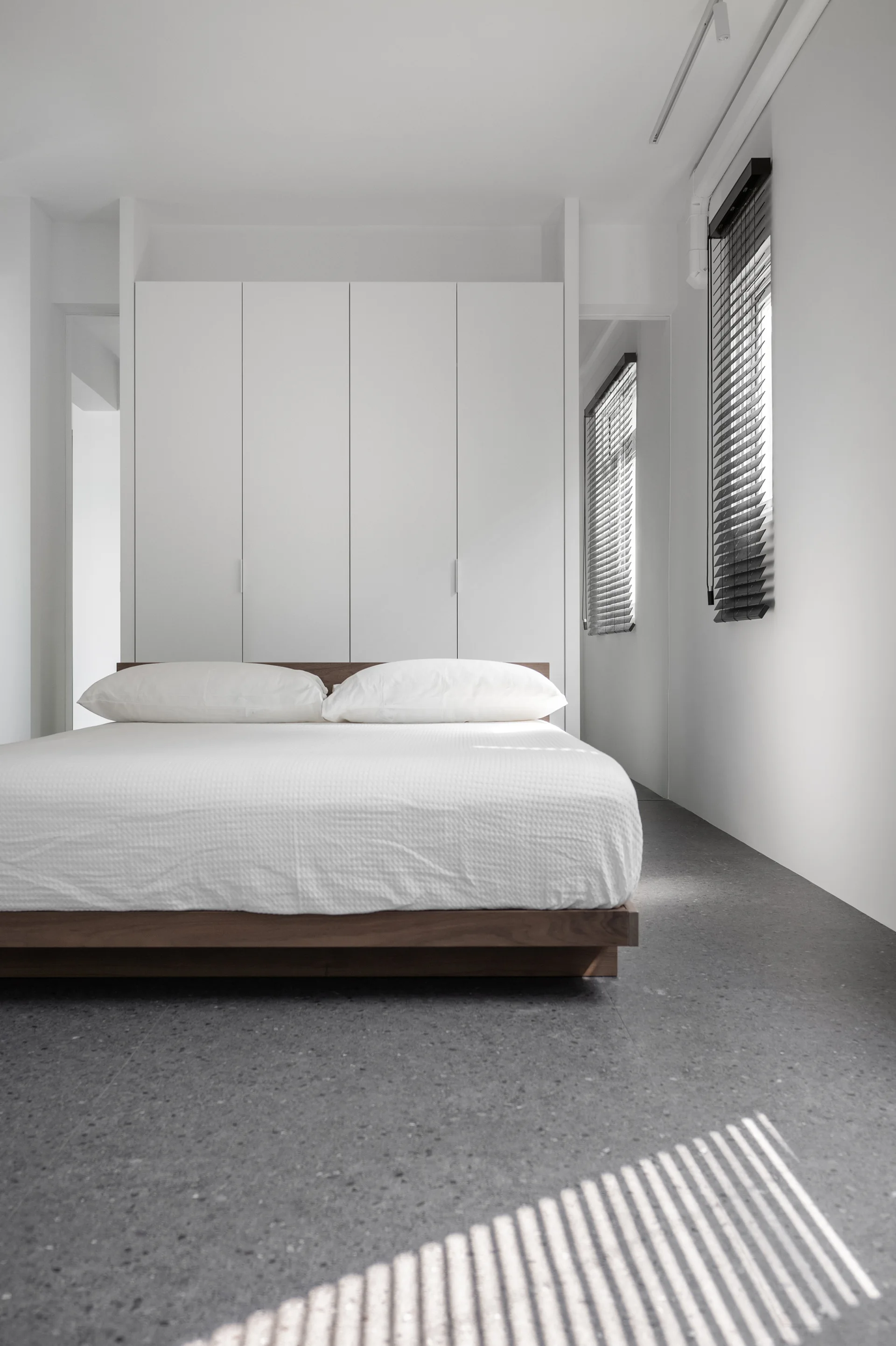 A minimalist bedroom features a low wooden platform bed with white bedding set against a seamless white built-in wardrobe on dark grey tile floors.