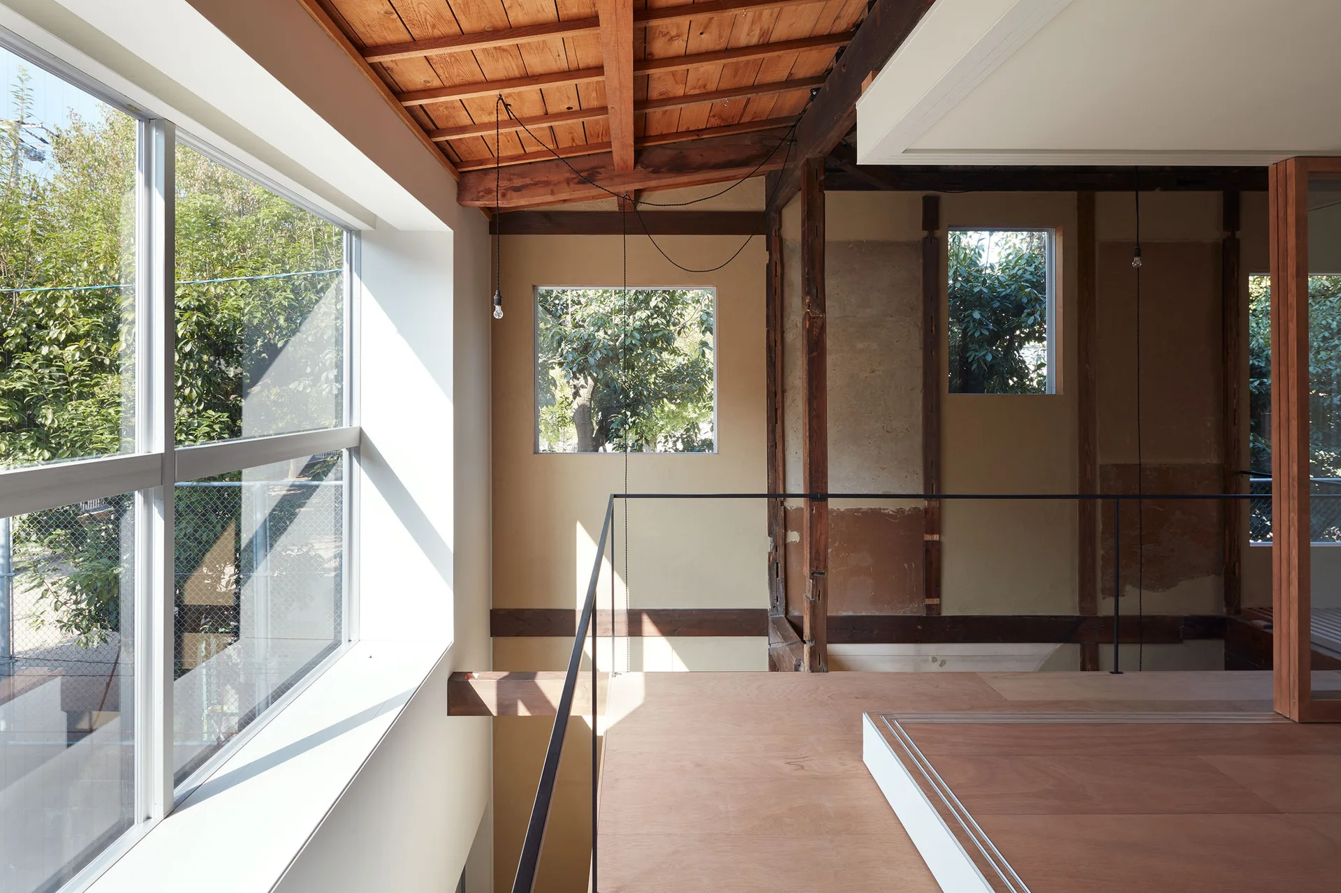 A modern mezzanine landing features exposed wood ceiling rafters, warm plaster walls, a thin black metal railing, and large windows looking out to trees.