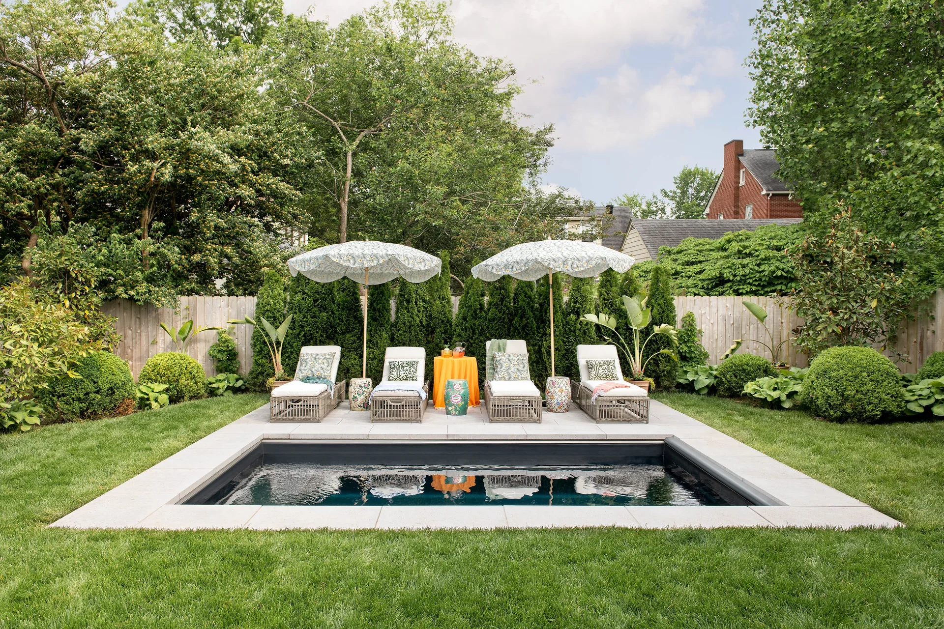 Four wicker chaise lounges sit on a stone patio by a rectangular swimming pool, flanked by floral-patterned umbrellas and a green hedge.
