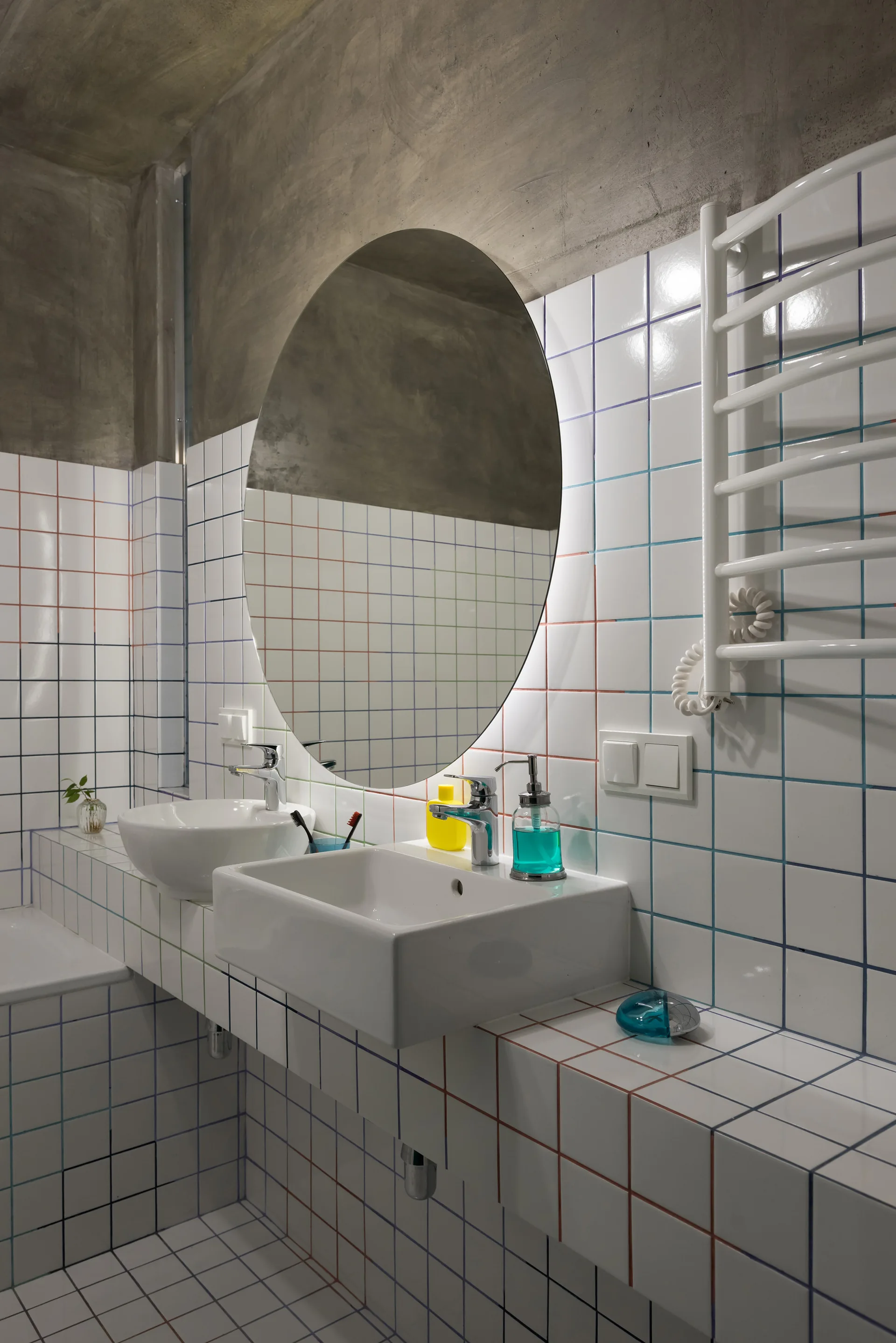 A modern bathroom features a tiled vanity with two sinks and an oval mirror against square wall tiles with multi-colored grout.