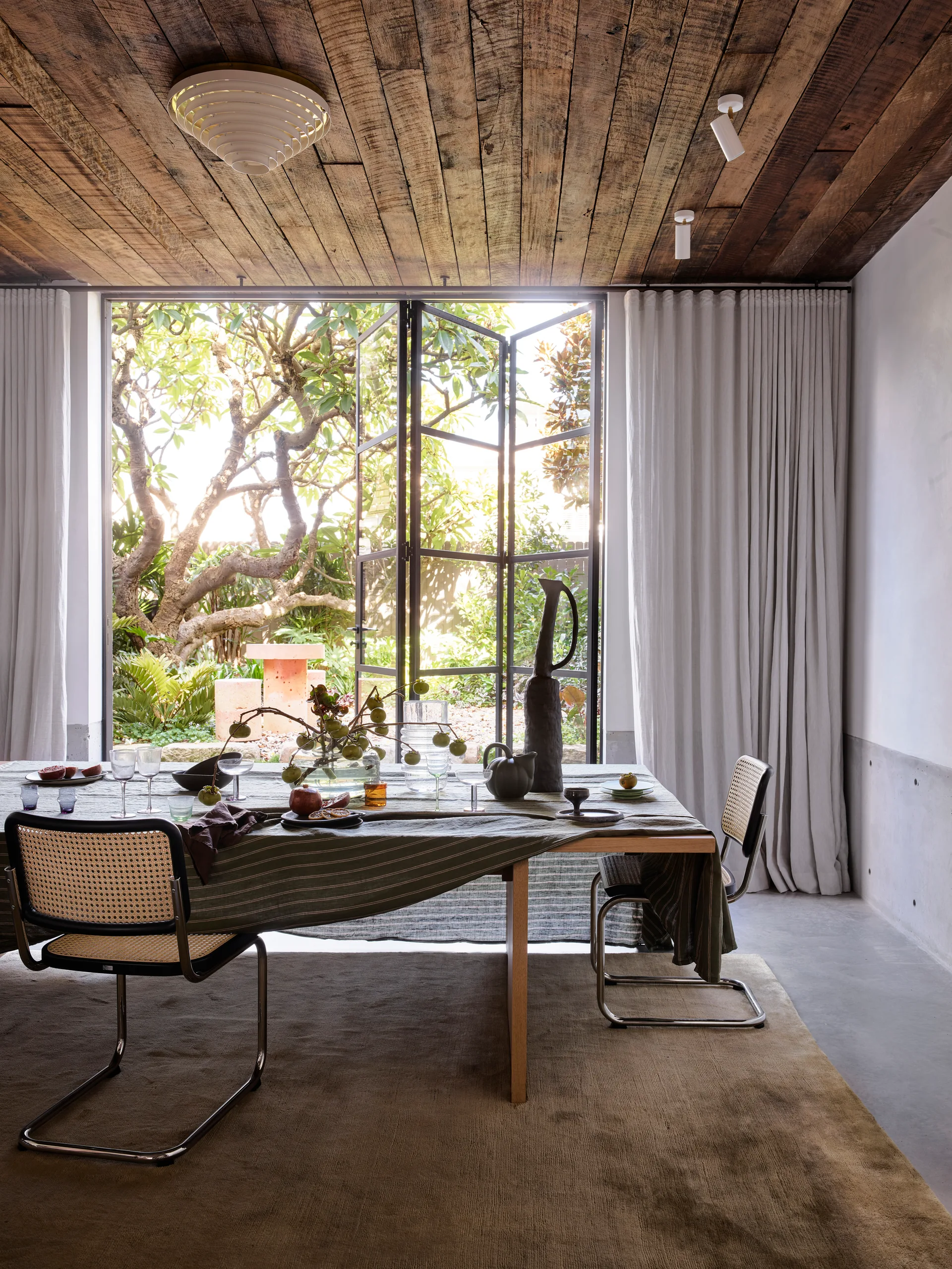 A modern dining room features a wood-topped table with cane chairs under a wood plank ceiling next to metal-framed glass doors that open to a garden.