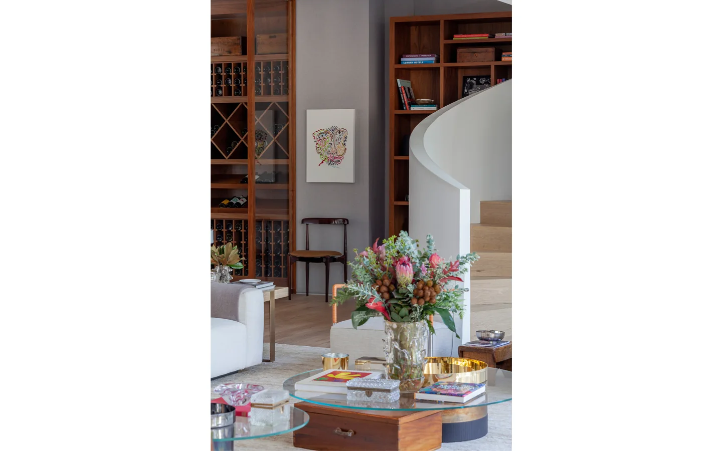 A contemporary living room featuring a curved white staircase, built-in wood shelving, a wine storage cabinet, and a glass coffee table.