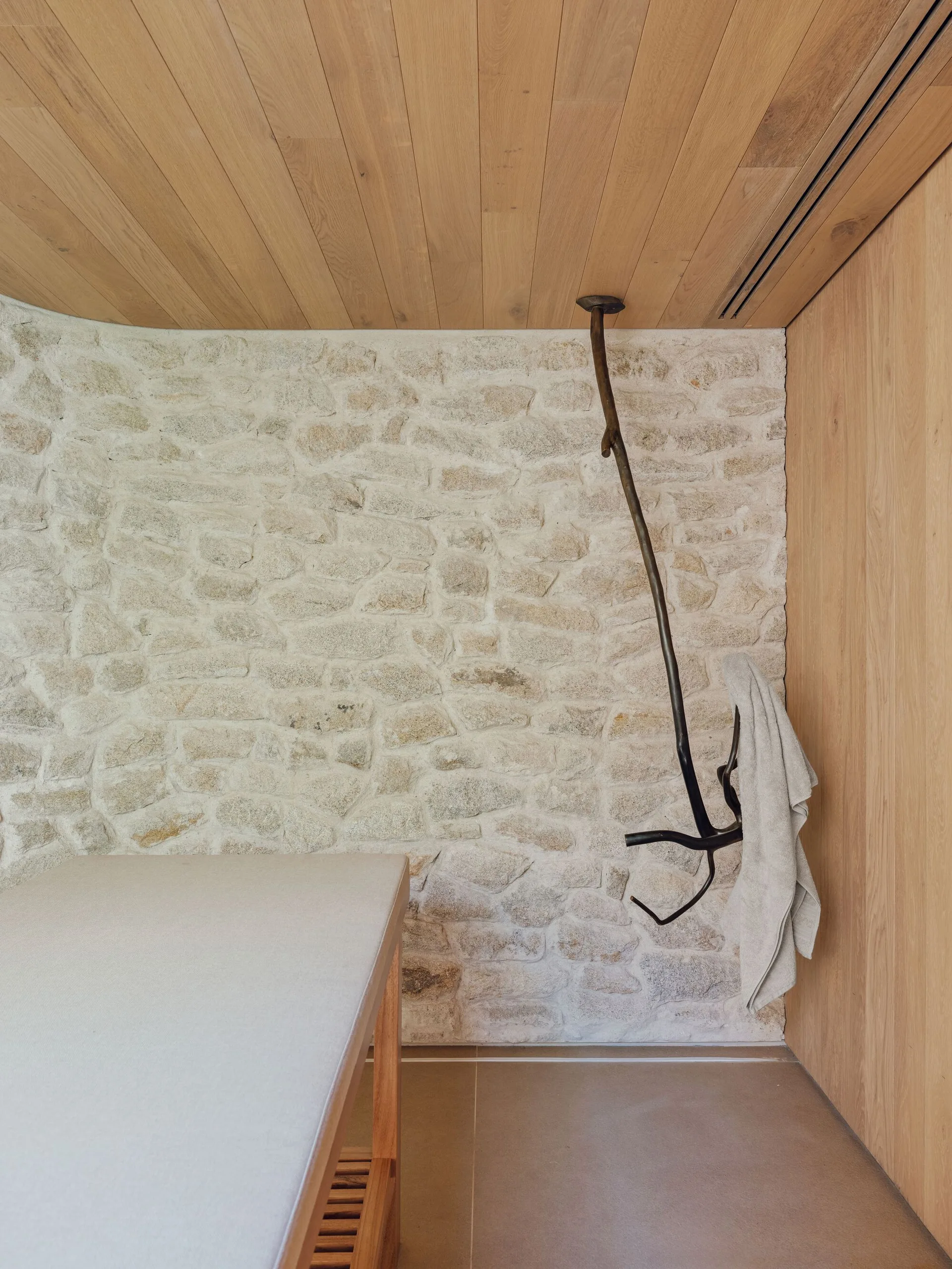 A minimalist wellness room featuring a wooden treatment table, a natural stone feature wall, a wood-paneled ceiling, and a branch-shaped hanging towel hook.