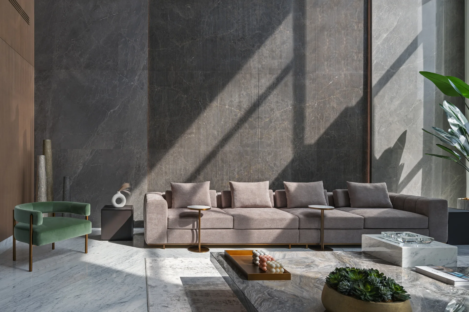 A contemporary living room featuring a long mauve sofa, a green velvet armchair, a fluted dark stone wall panel, and polished marble flooring.