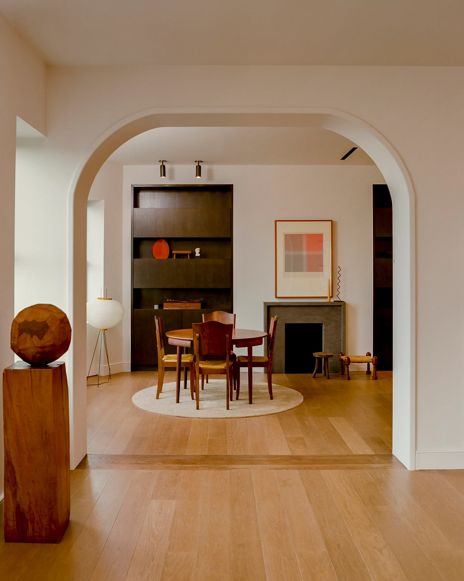 A minimalist dining room with a round wooden table and chairs is visible through a large, smooth plaster archway under soft lighting.