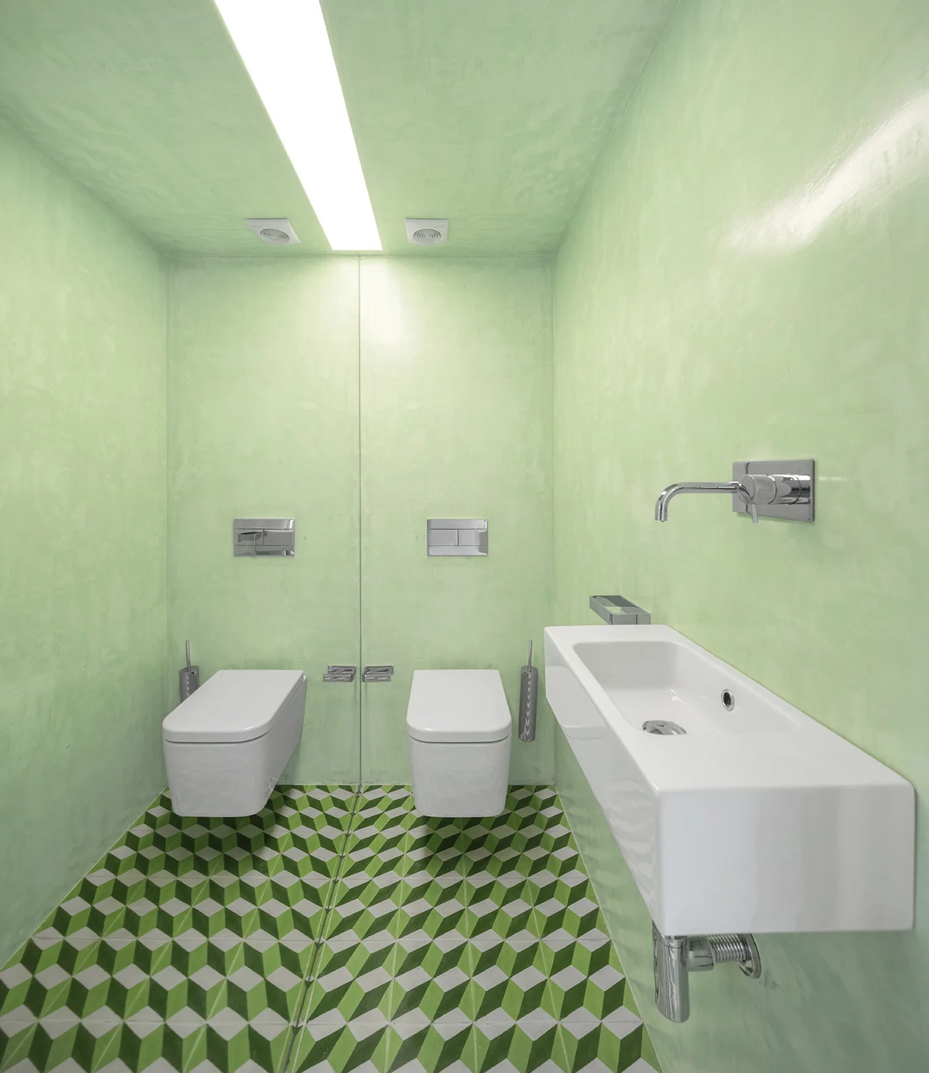 A minimalist green powder room features textured plaster walls, a wall-mounted white trough sink, and green geometric floor tiles.