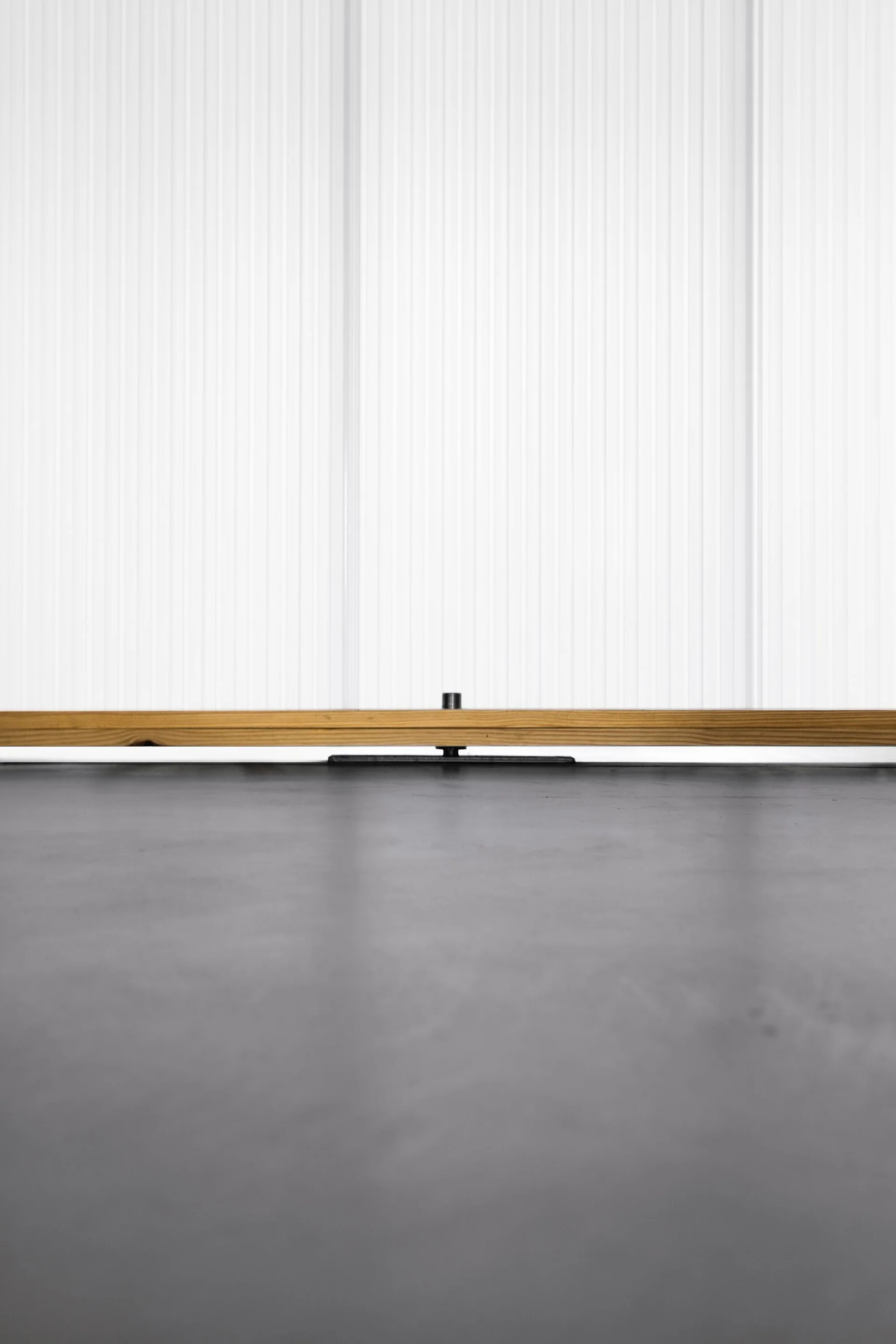 A close-up architectural detail showing a white vertically ribbed wall panel meeting a smooth grey concrete floor with a natural wood baseboard.