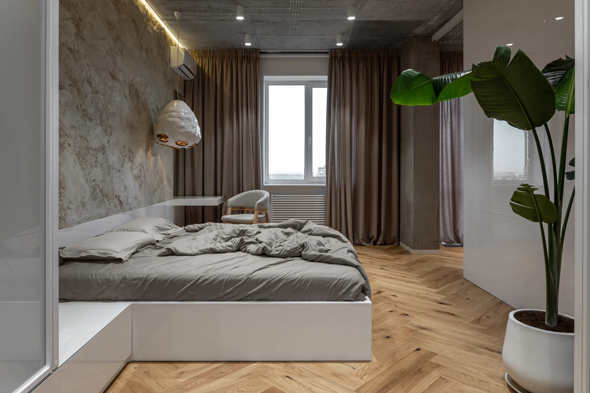 A minimalist bedroom features a low-profile bed with grey linens, herringbone hardwood flooring, an exposed concrete ceiling, and brown drapes.
