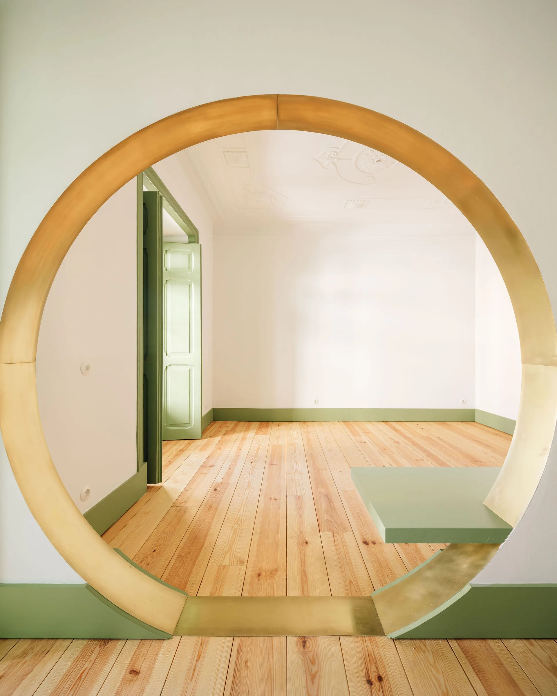 A circular brass archway frames an empty white room featuring sage green baseboards, a matching open door, and light hardwood floors.