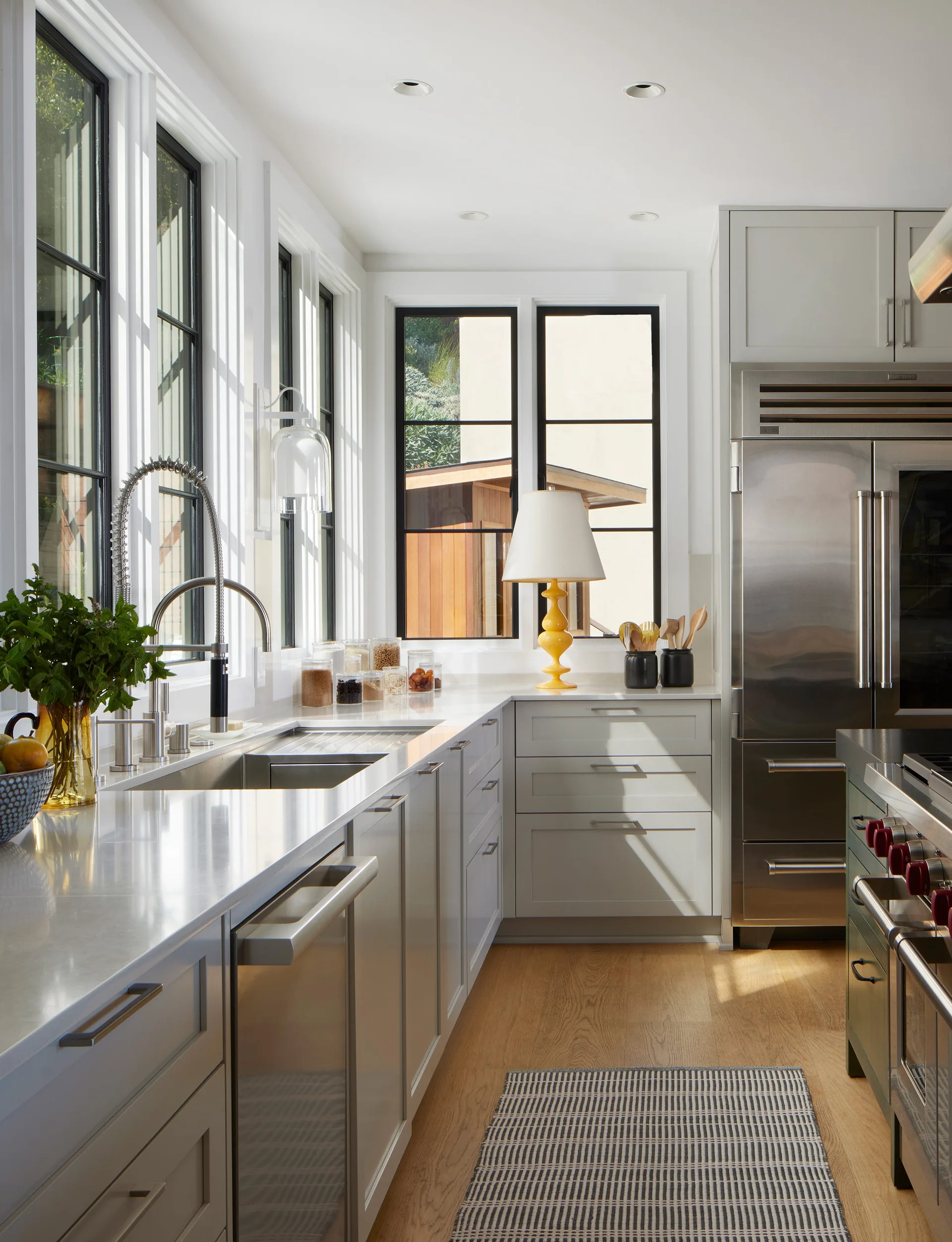 A light gray kitchen features black-framed windows, stainless steel appliances, and a bright yellow table lamp on the countertop next to a sink.