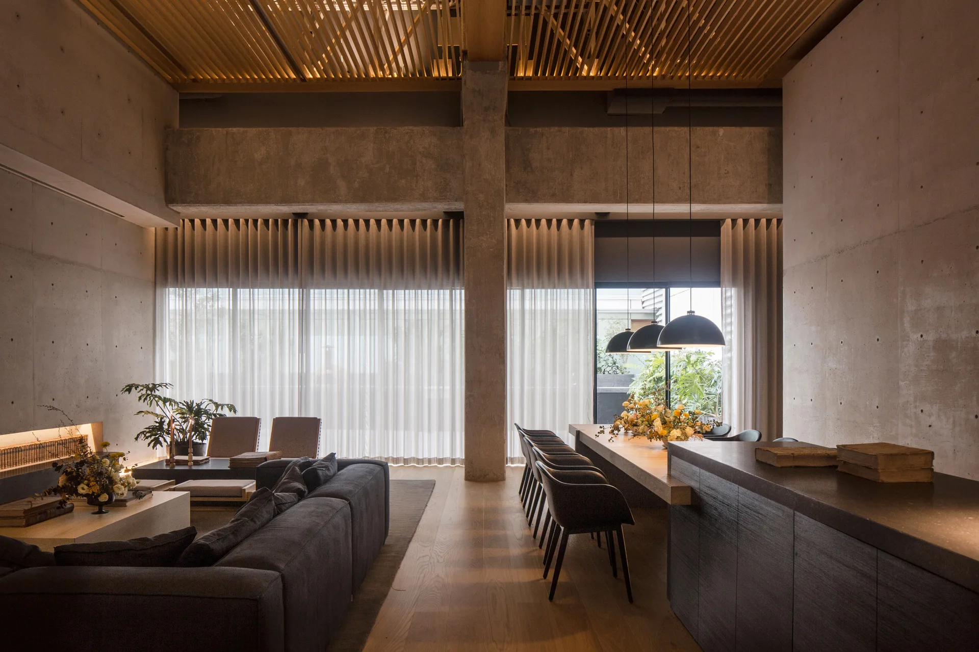 An open-concept living and dining area featuring textured concrete walls, a slatted wood ceiling, a grey sectional sofa, and a long dining table.