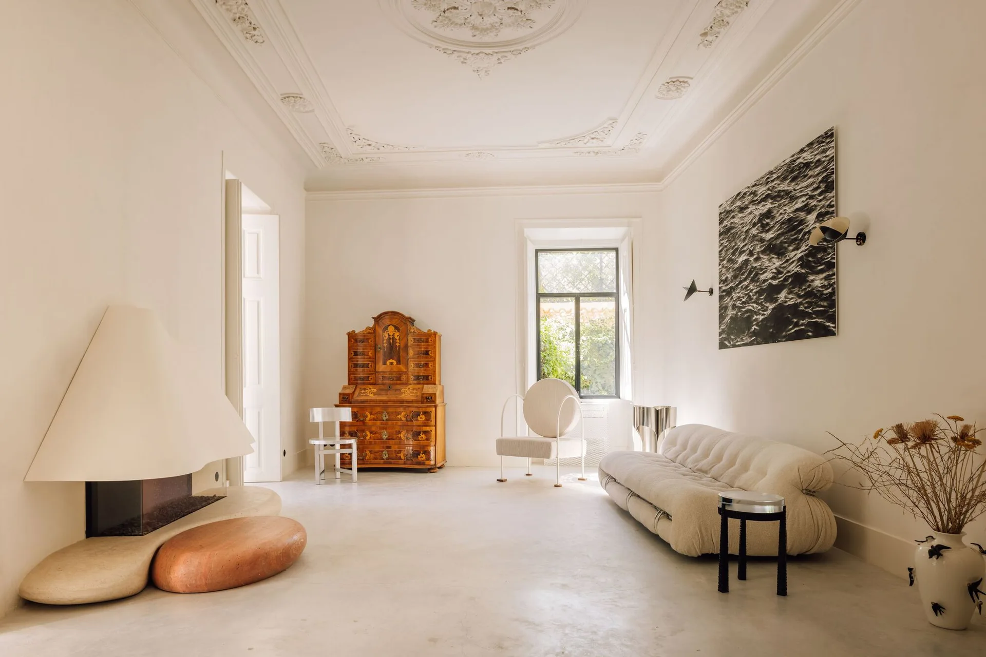 A minimalist living room featuring a conical white fireplace, a low-profile cream sofa, and an antique wooden cabinet under an ornate plaster ceiling.