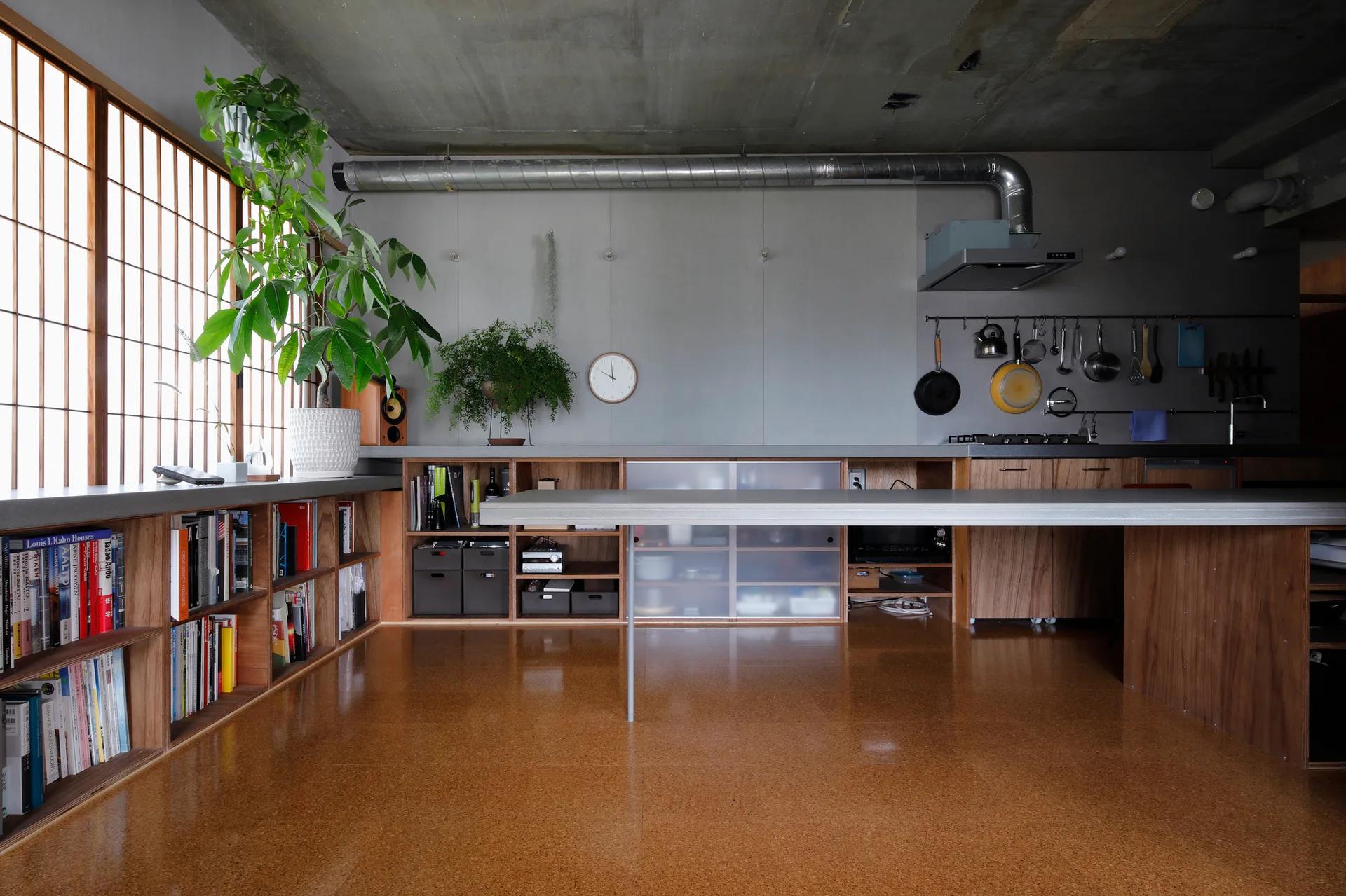 An open-concept kitchen features a long grey countertop, low wooden cabinetry, a shoji screen window wall, and exposed metal ductwork.