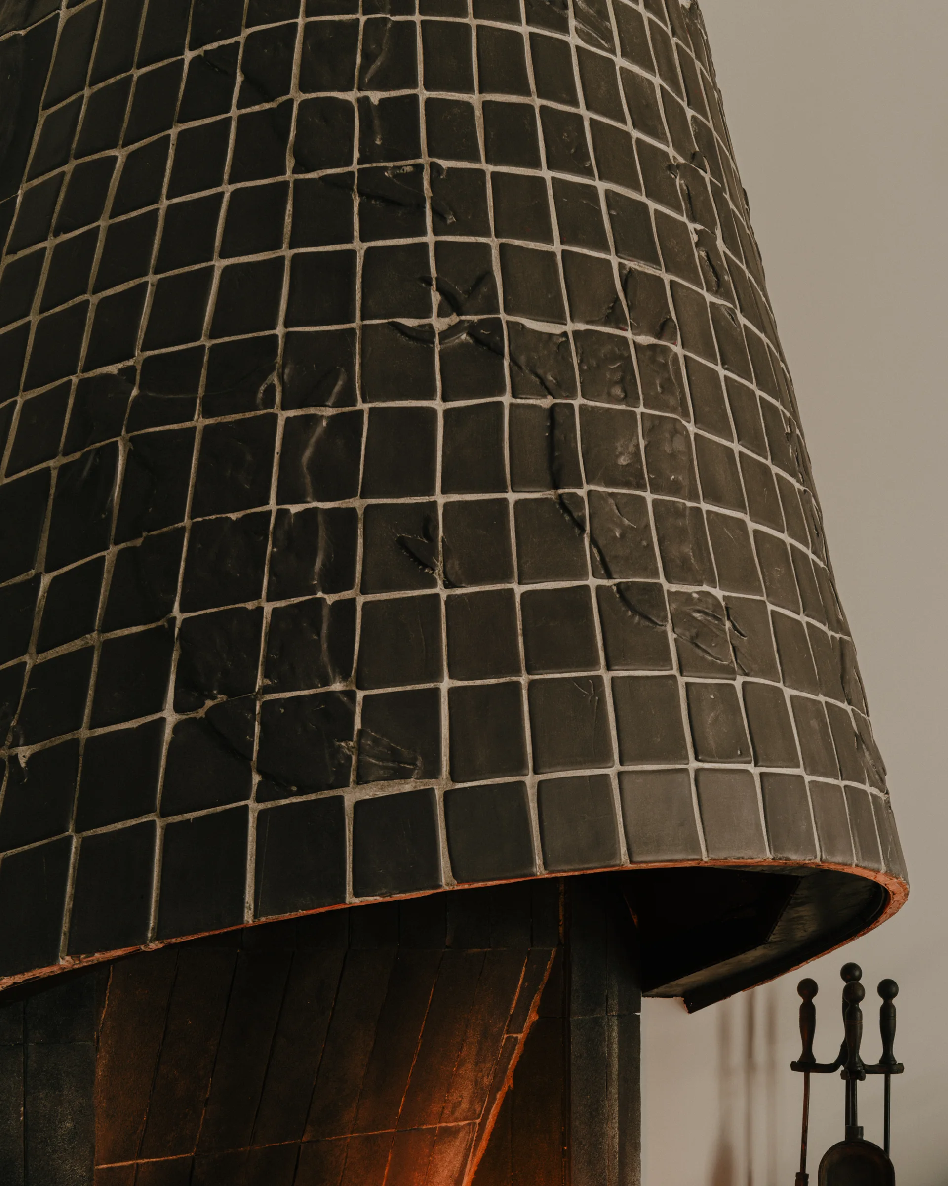 A close-up of a curved fireplace hood clad in dark charcoal square ceramic tiles with a textured finish against a light wall.
