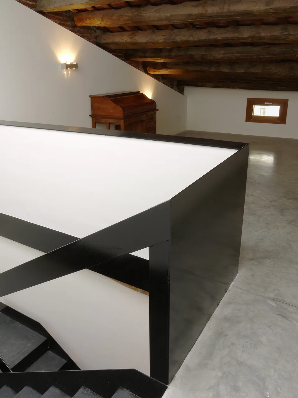 A black minimalist staircase railing sits on a polished grey concrete floor beneath rustic exposed wooden ceiling beams and a white wall.