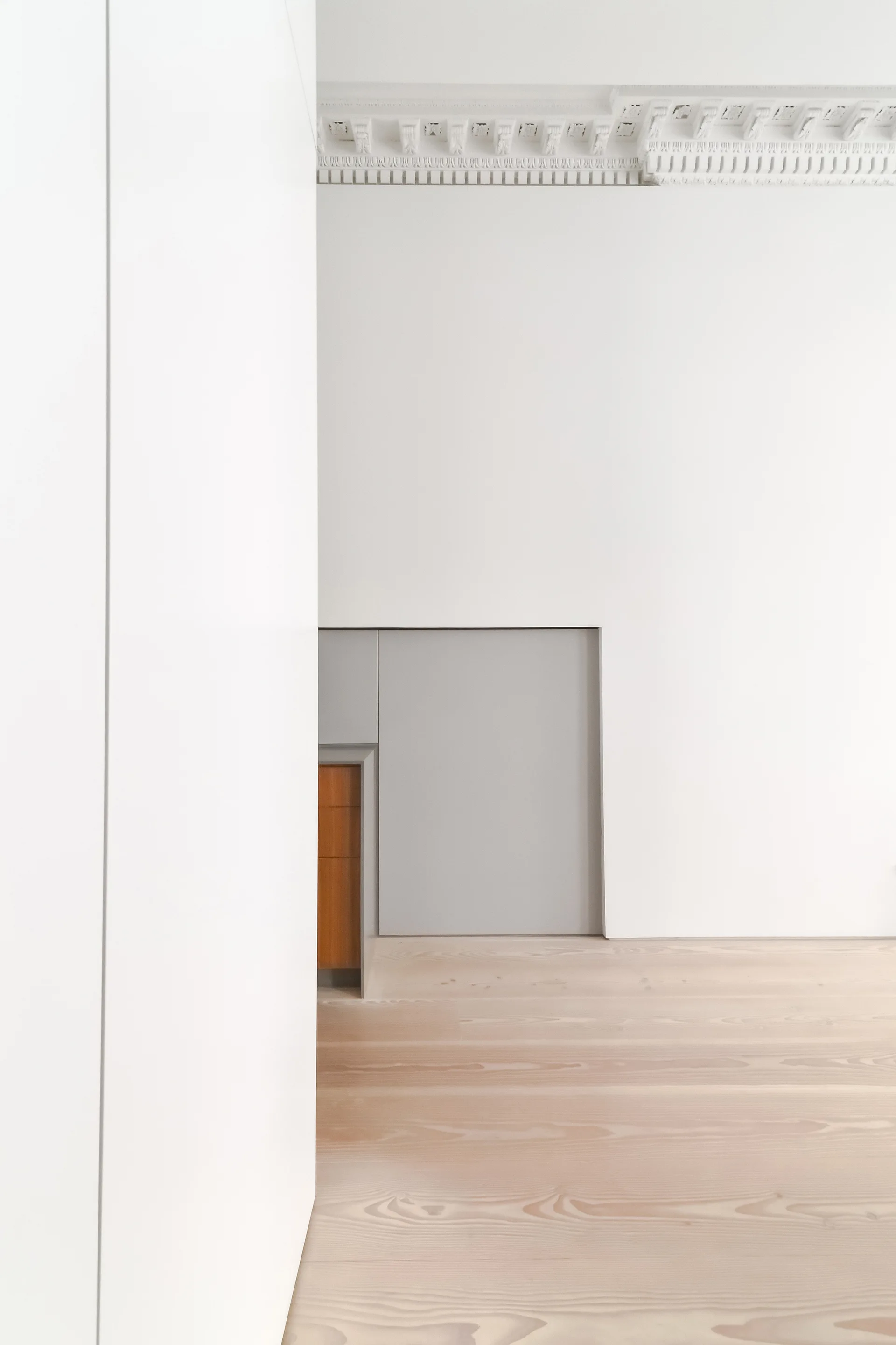 A minimalist white room with detailed ceiling crown molding, light wood plank floors, and a recessed storage cabinet built into the wall.
