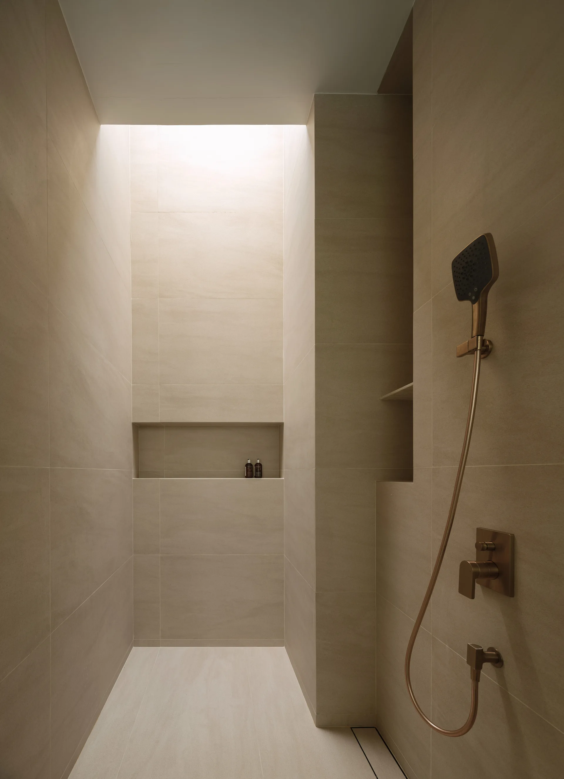 A minimalist walk-in shower features beige tiled walls, a rectangular built-in niche, bronze fixtures, and natural light from a ceiling skylight.