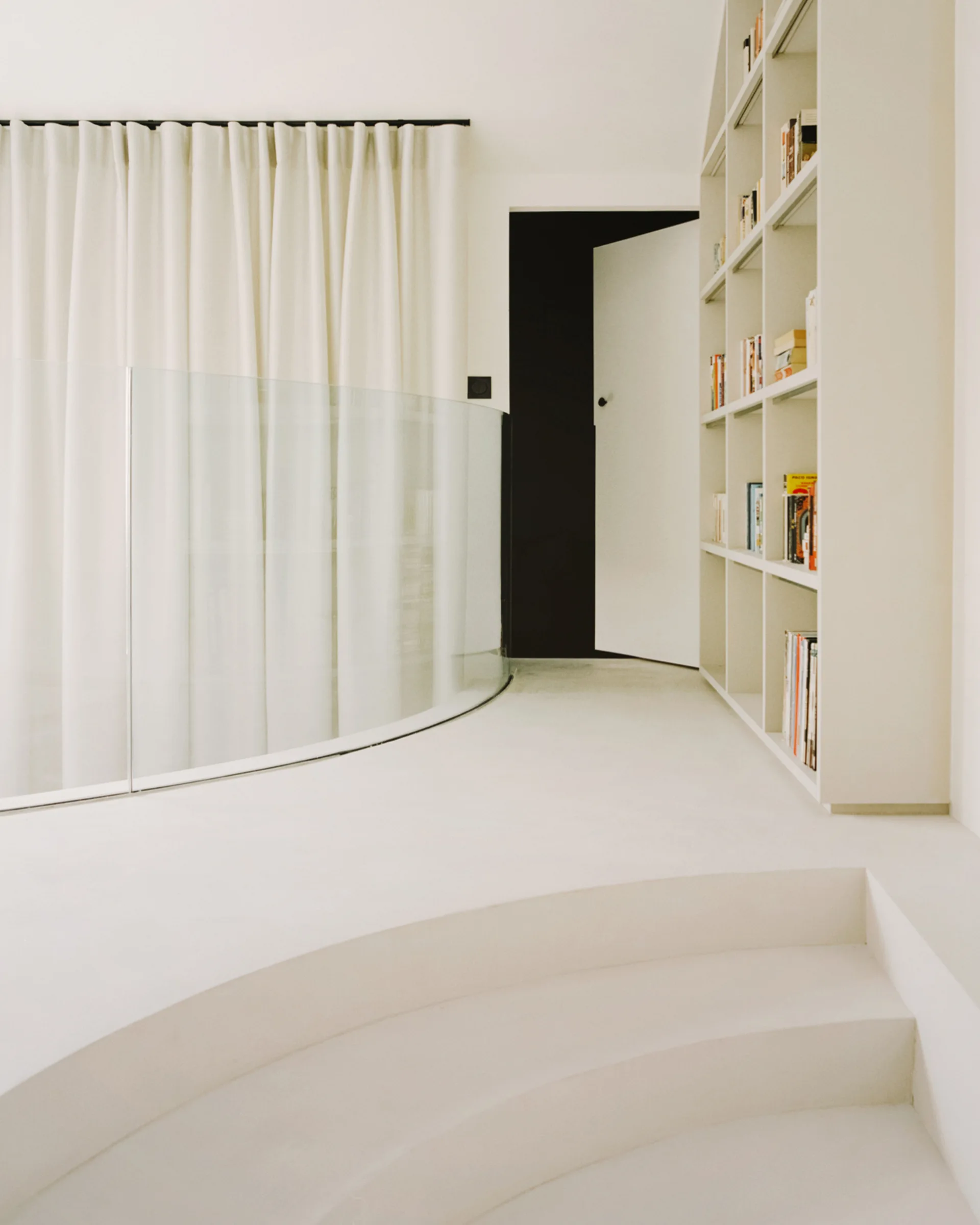 A minimalist white staircase landing features a curved glass balustrade, floor-to-ceiling white drapes, and a built-in bookshelf.