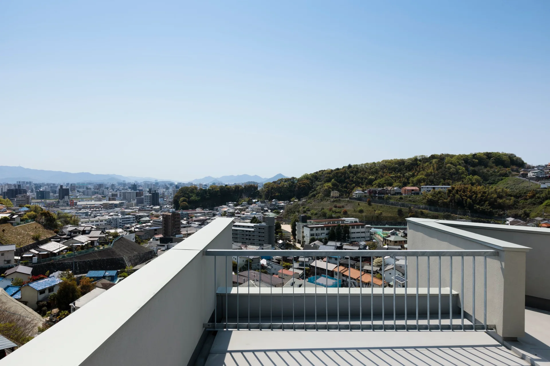 An elevated concrete rooftop terrace featuring a silver metal railing and solid parapet walls that overlook a sprawling hillside city under a clear sky.