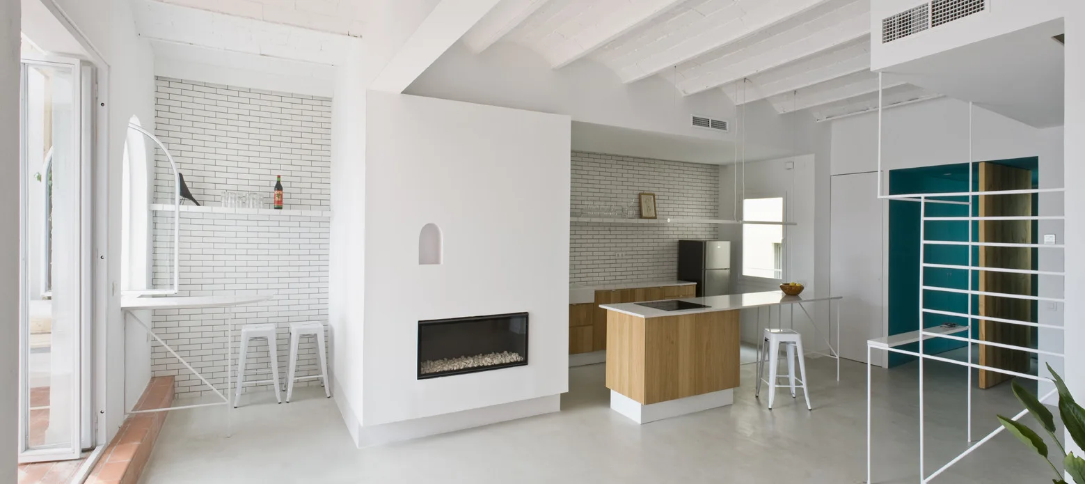 A minimalist open-plan white kitchen features a wood-clad island, white brick walls, exposed ceiling beams, and a modern fireplace partition.