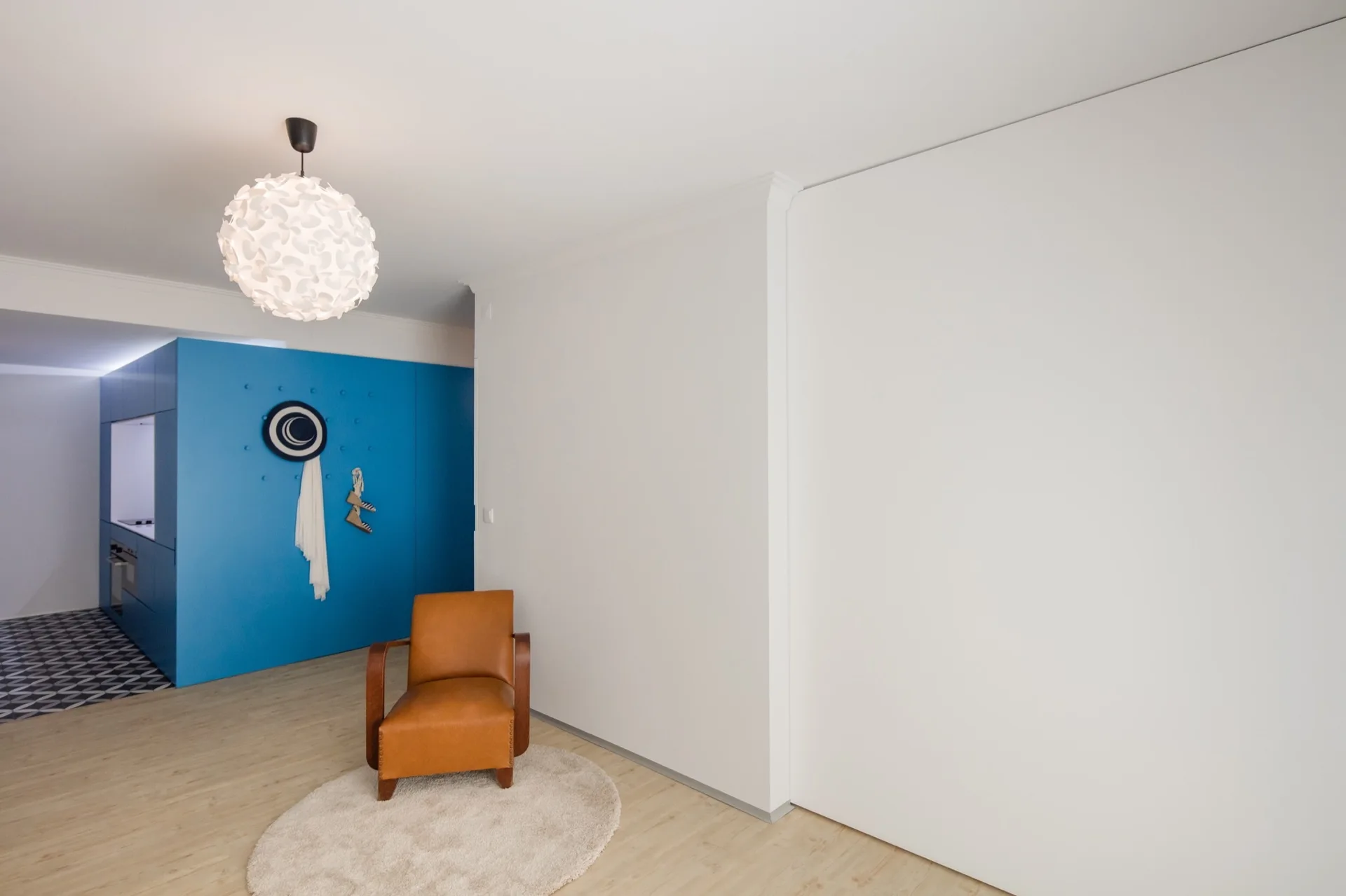 A tan leather armchair rests on a round cream rug in a minimalist white room featuring a bright blue storage unit and a globe pendant light.